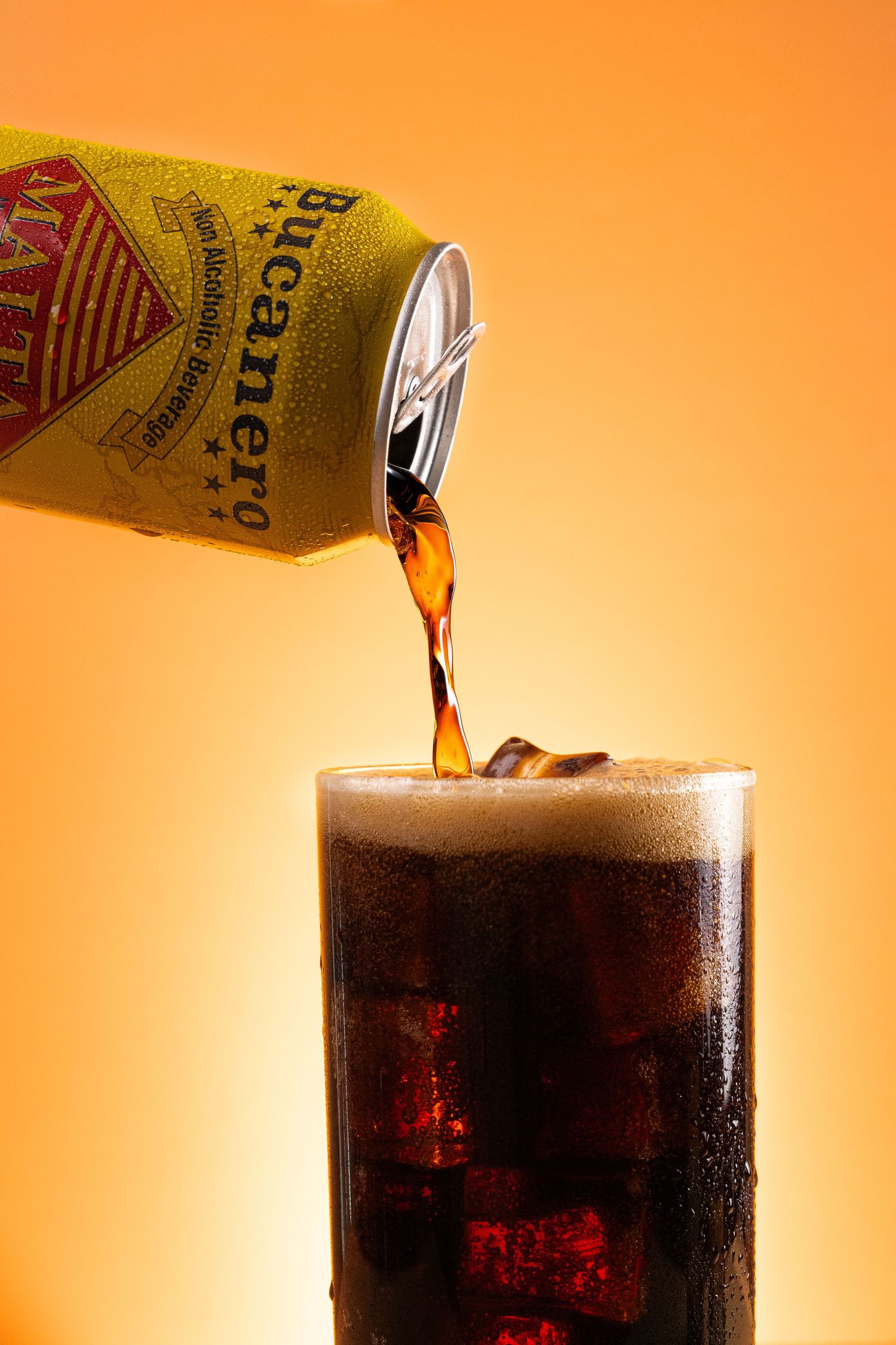 Carbonated cola being poured from a yellow can into a glass filled with dark soda and ice cubes against an orange background.