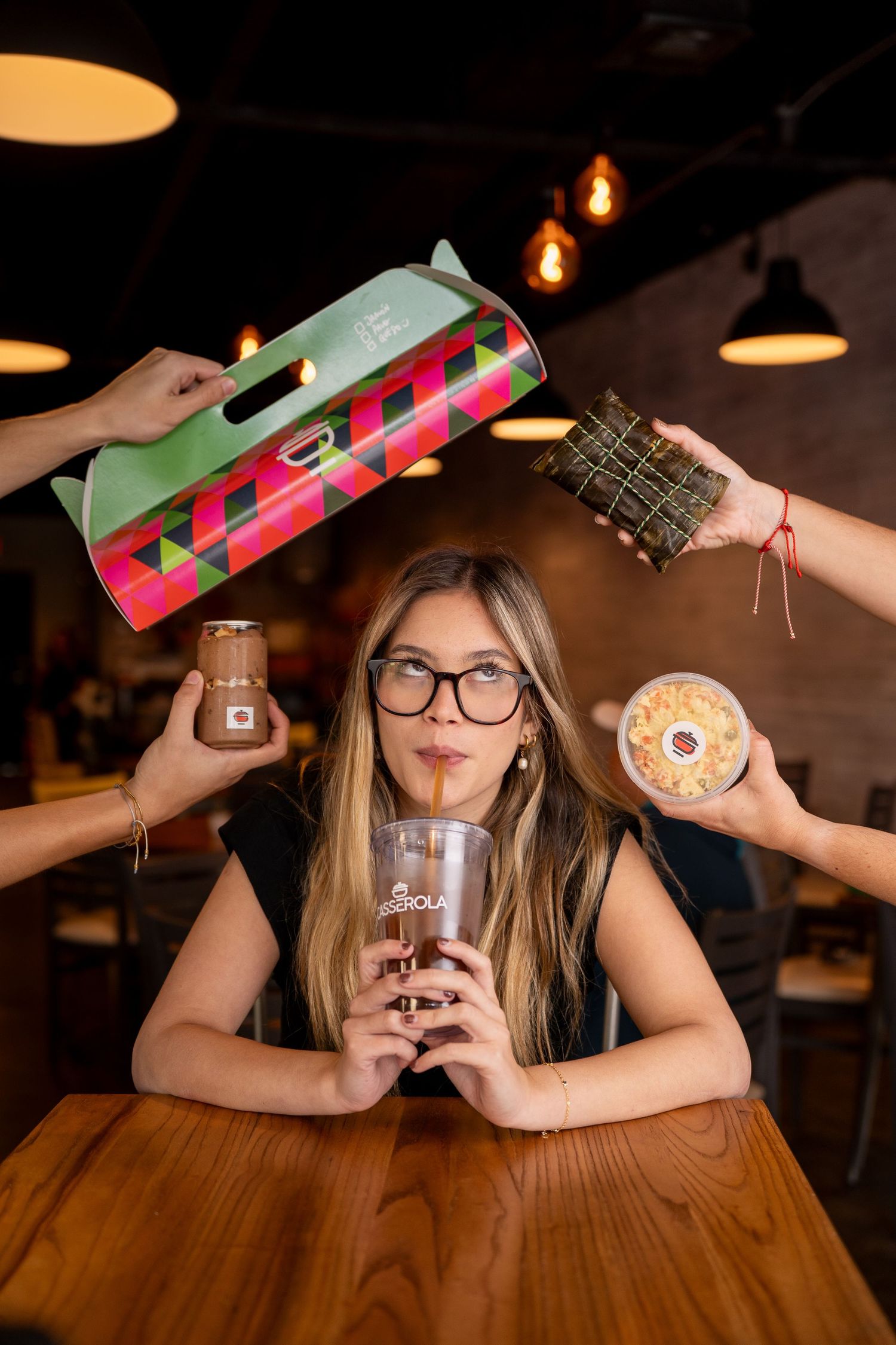 Woman in glasses sips iced coffee at cafe table while hands offer colorful box, tamale, jar, and pastry.