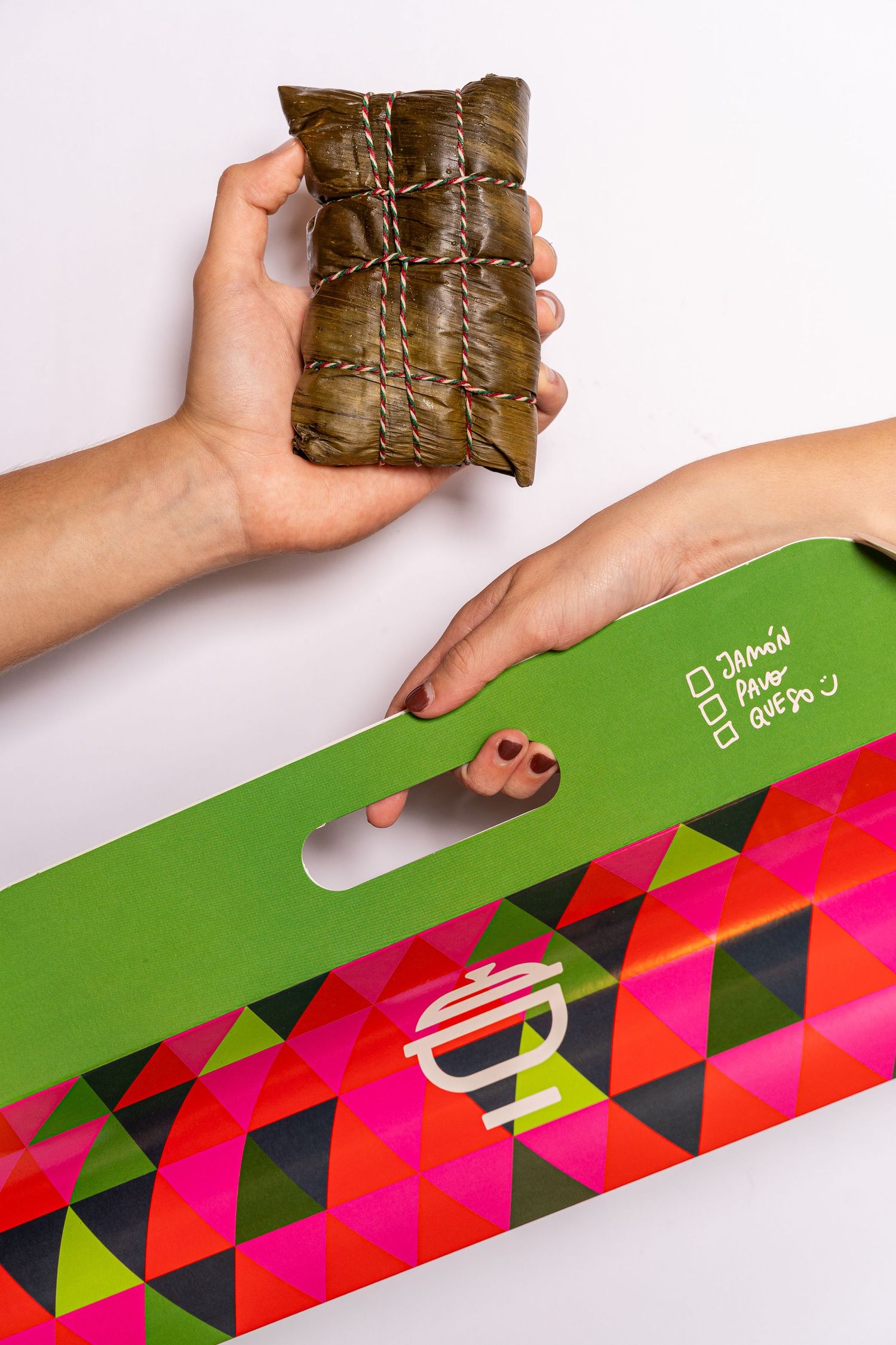 Hands holding a traditional tamale wrapped in banana leaves above a colorful green and pink geometric patterned box.
