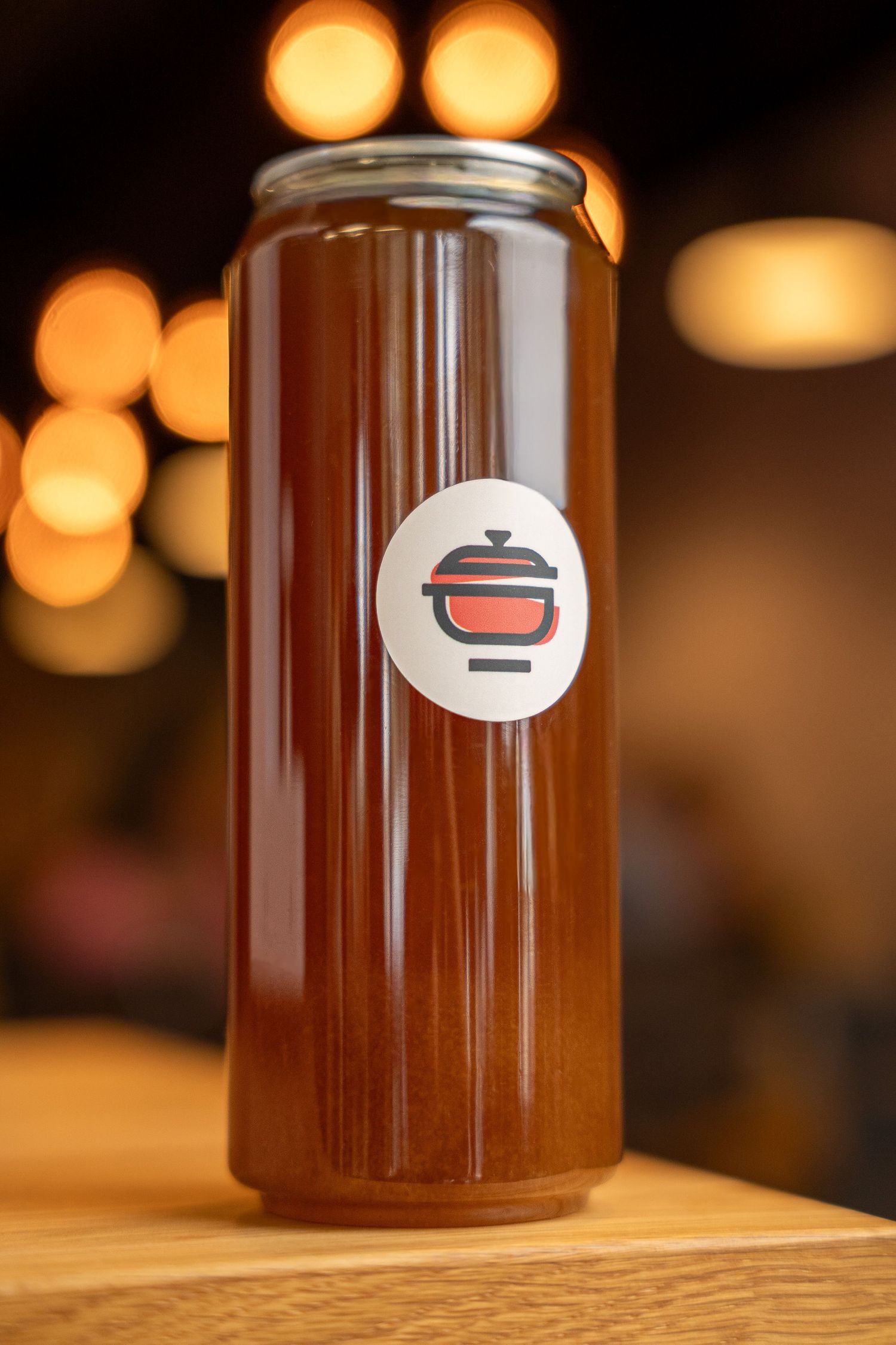 A tall glass can filled with amber liquid, featuring a red pot logo sticker, set on a wooden surface with bokeh lights.