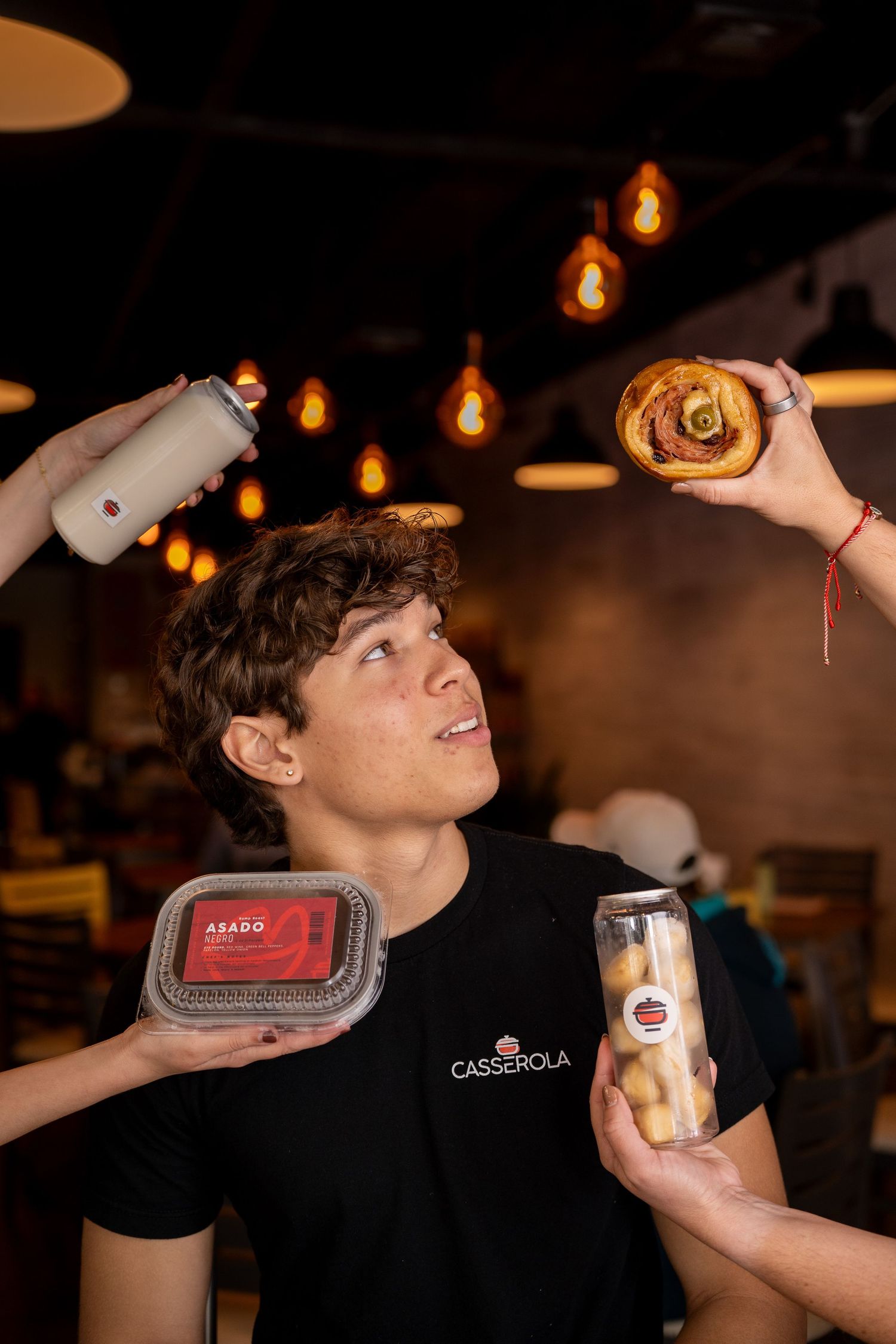 Young man in Casserola shirt surrounded by hands holding pastry, spray can, food container, and jar in a restaurant.