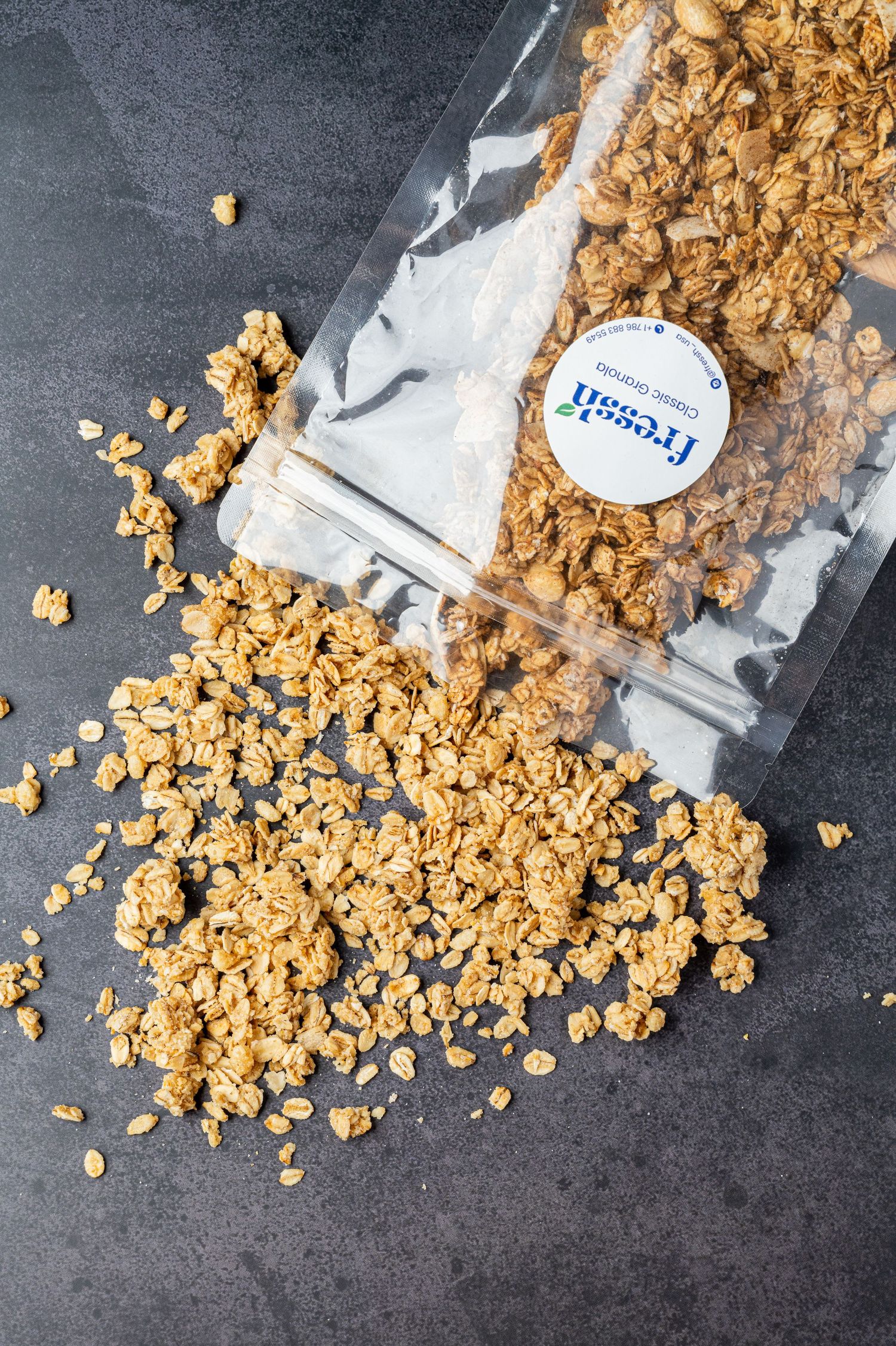 Granola spilling out of a Fresh Craft resealable bag onto a dark stone surface.