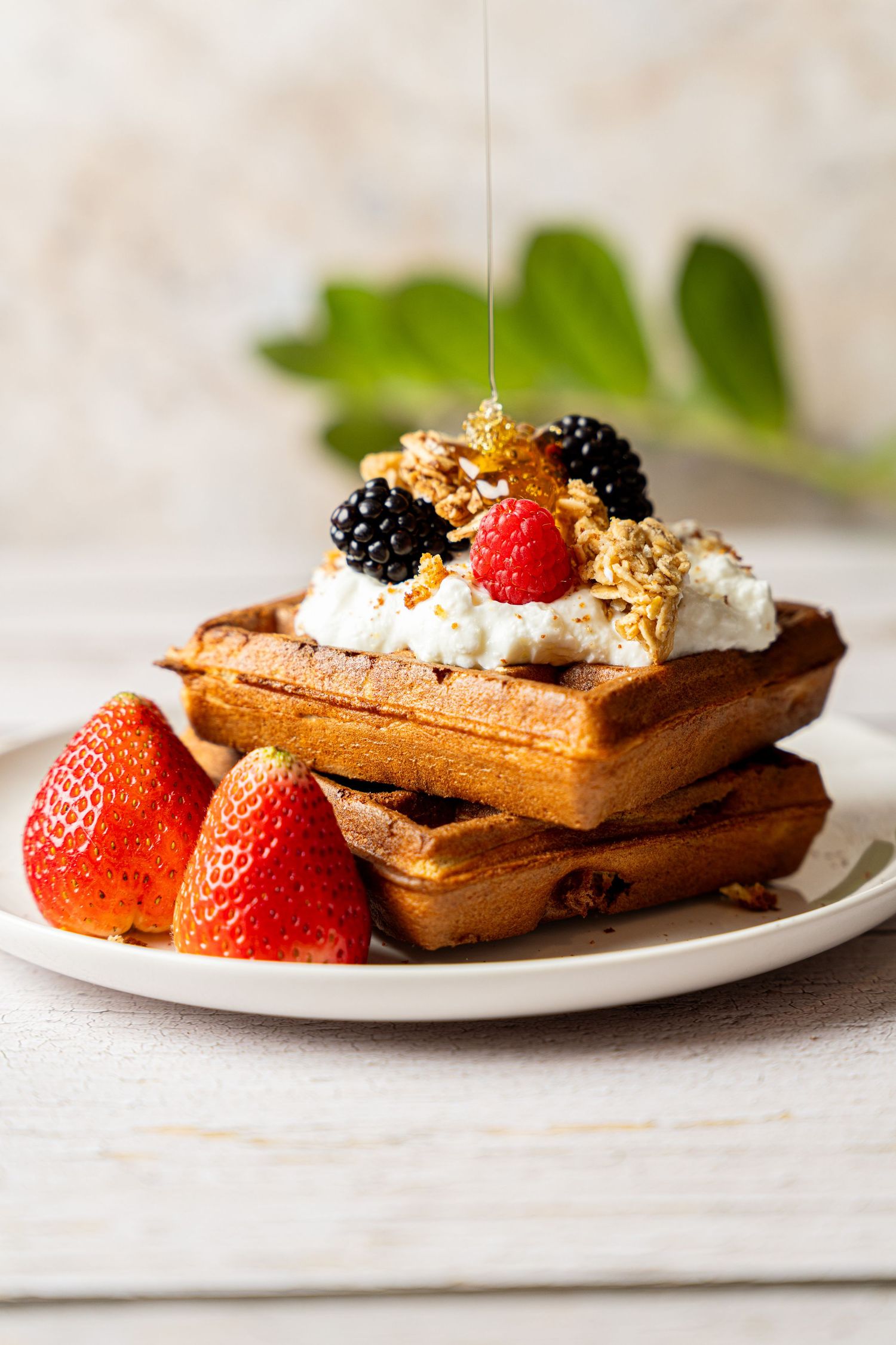 Stack of golden waffles topped with whipped cream, blackberries, raspberry, granola, and honey, served with fresh strawberries.