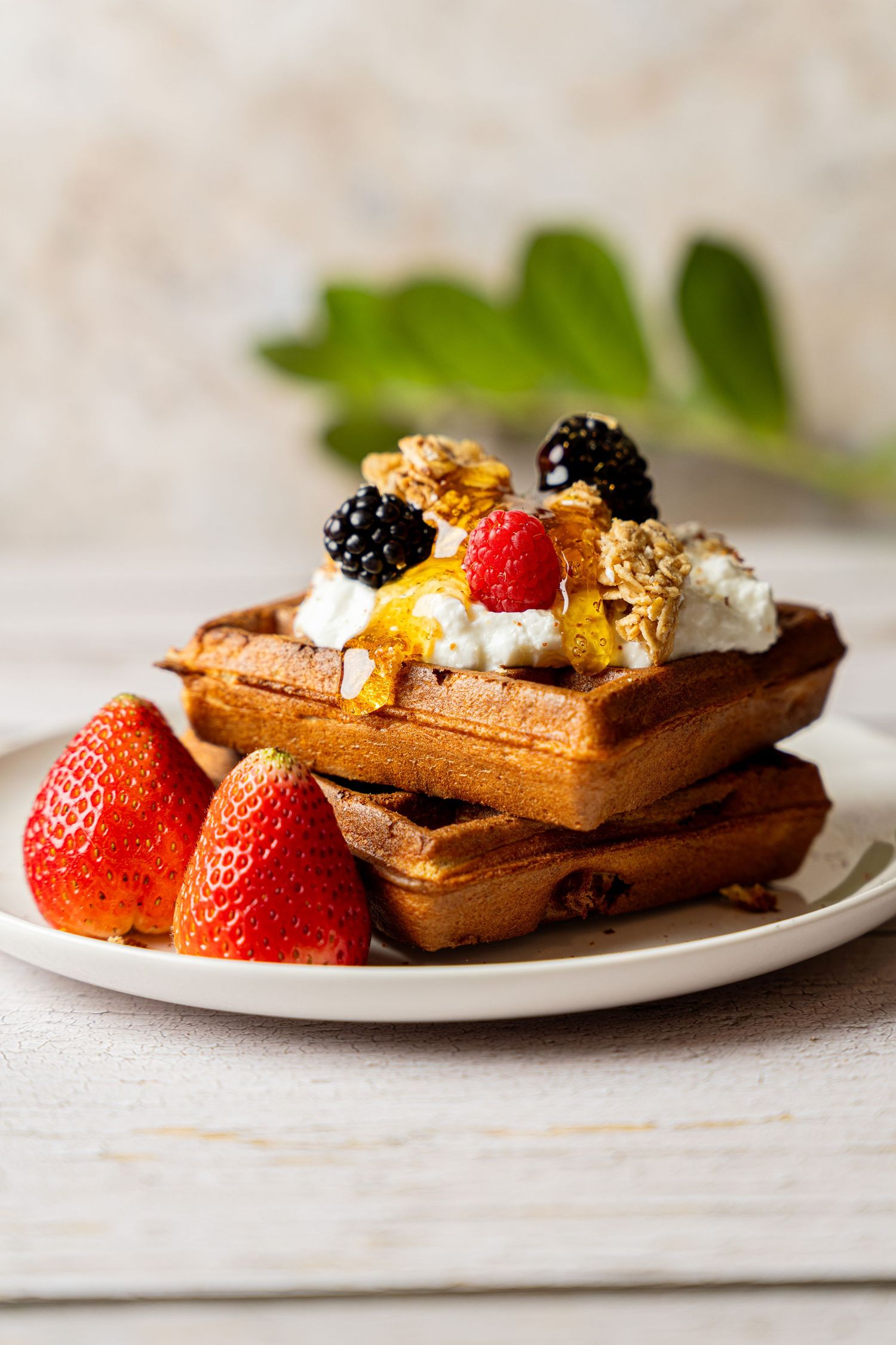Stack of golden waffles topped with whipped cream, granola, blackberries, and strawberries on a white plate.
