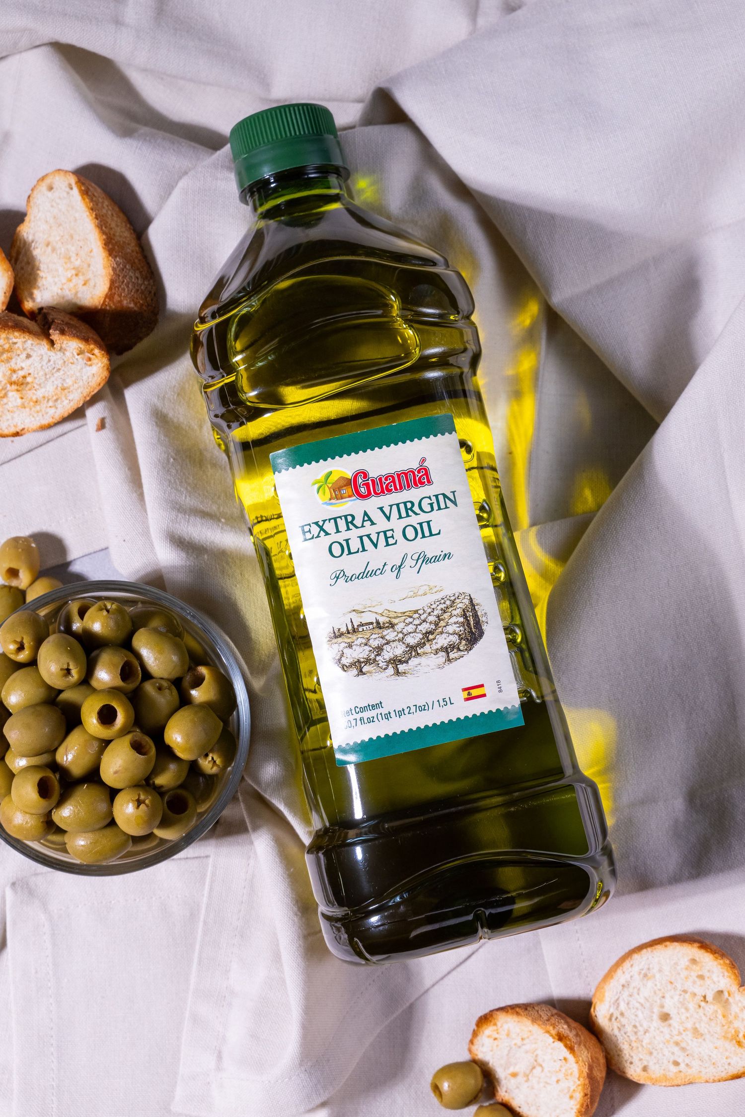 Bottles of extra virgin olive oil displayed with green olives and bread slices on white linen tablecloth.