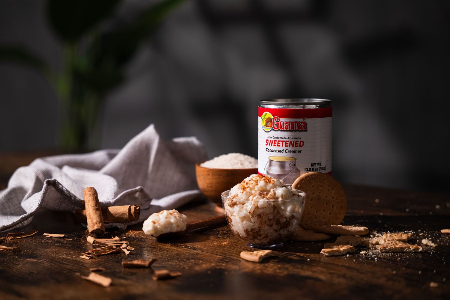 Sweetened condensed milk can with coconut flakes, cinnamon sticks, and brown sugar on a rustic wooden surface.