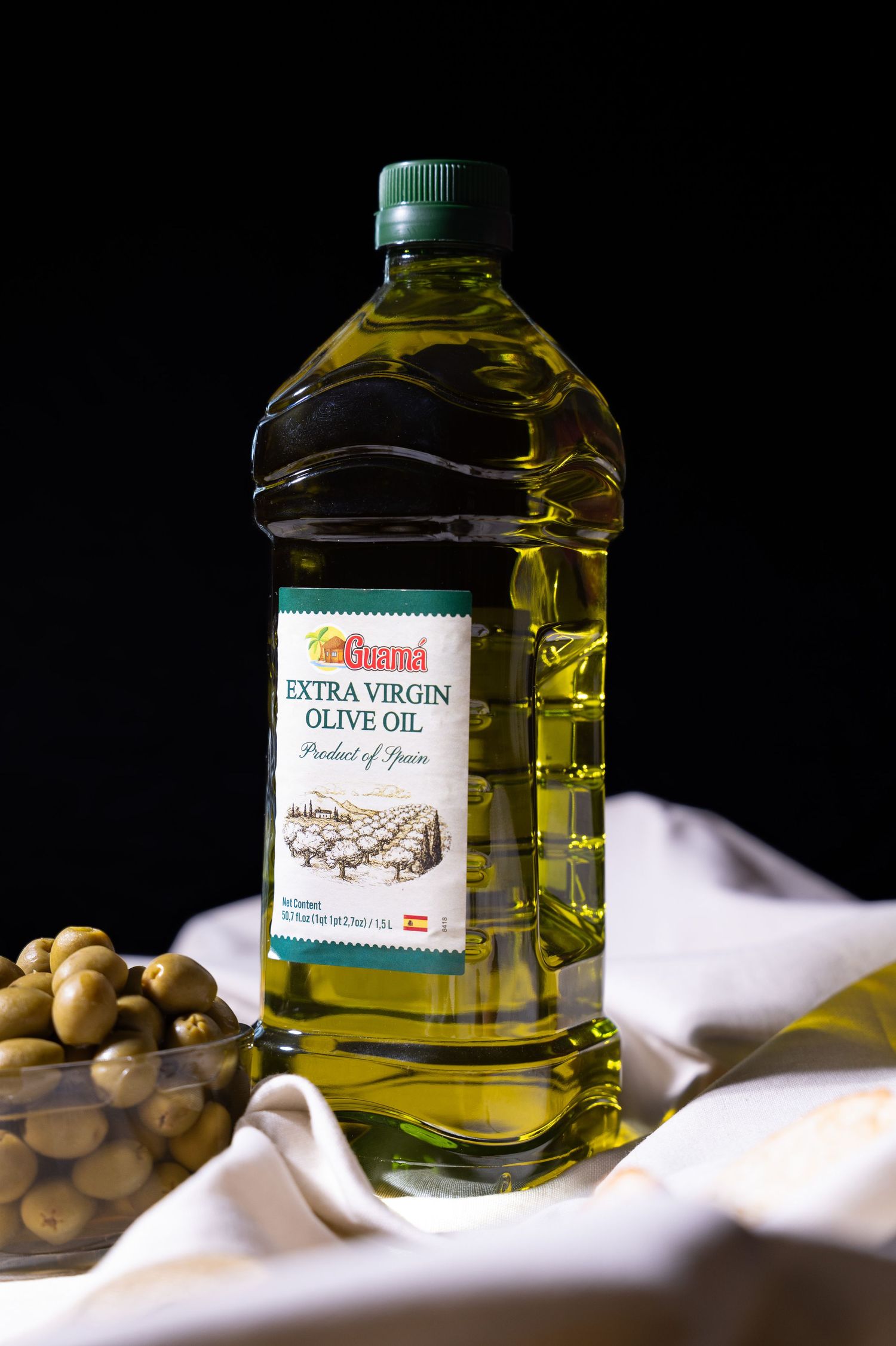 A bottle of extra virgin olive oil sits beside green olives on a white cloth with dramatic lighting and shadows.