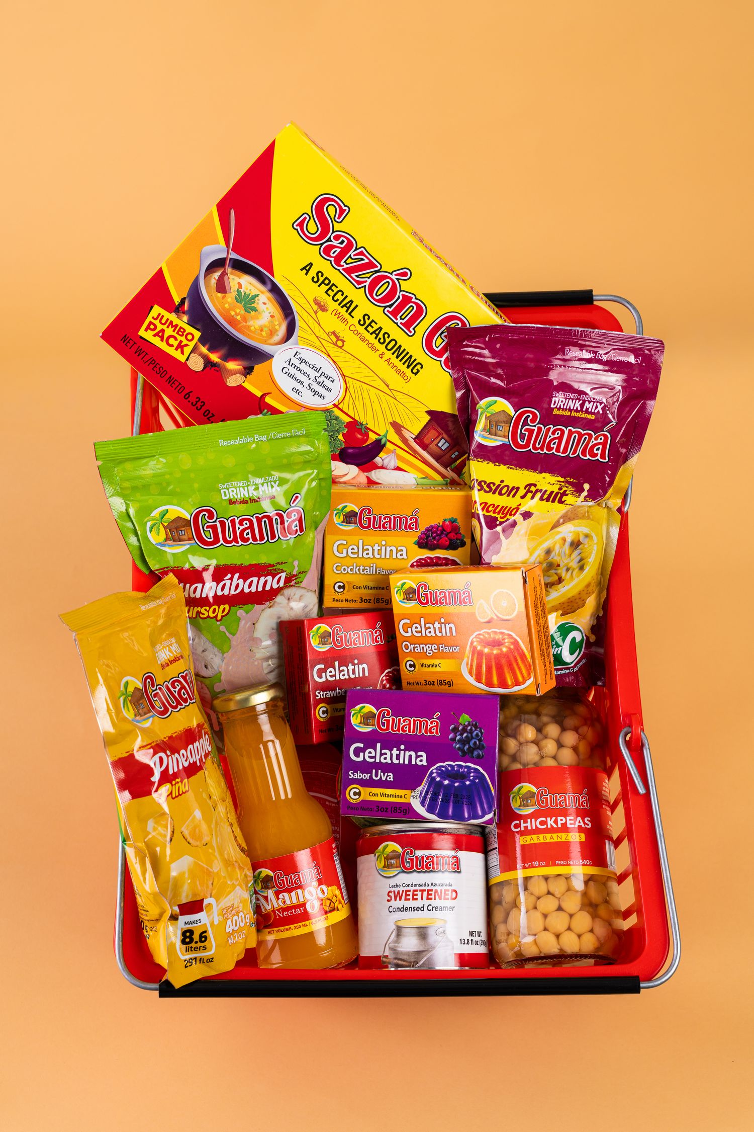 Red shopping basket filled with Latin grocery items including Sazon, Guama gelatin, chickpeas, and fruit syrup products.