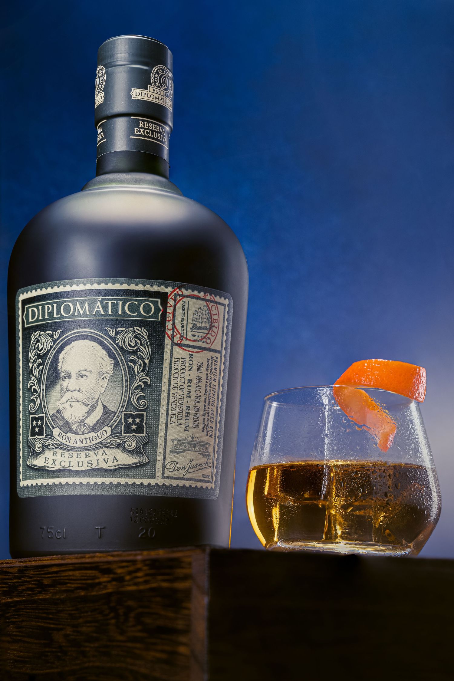 A bottle of Diplomatico rum next to a rocks glass with orange peel garnish against a dark blue background.