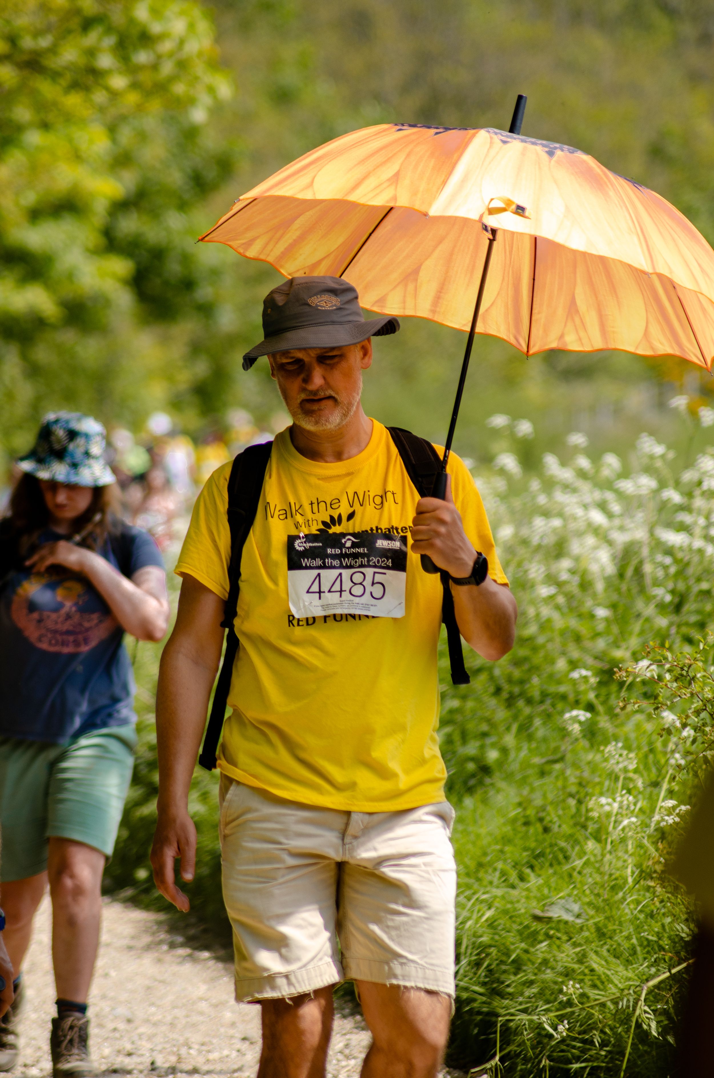 Participant with orange umbrella walks sunny trail wearing yellow event shirt and race number.