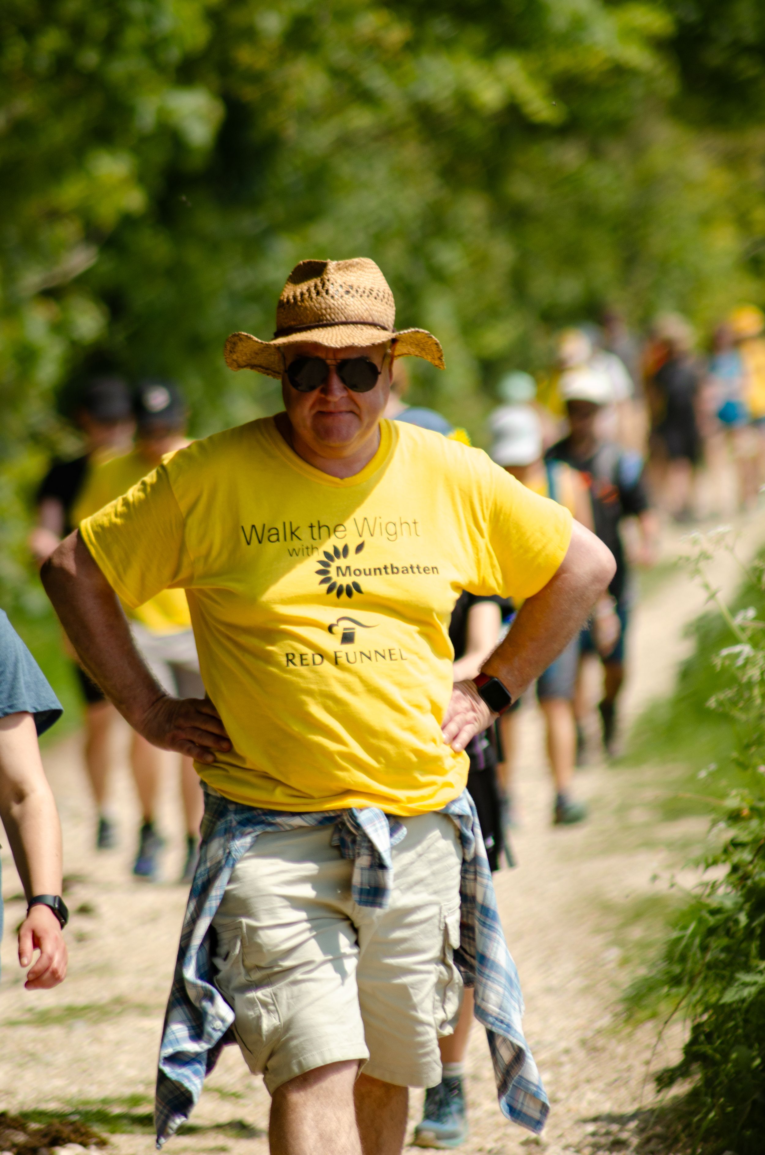 Participant wearing a yellow t-shirt and straw hat walking on a sunny outdoor trail with other people.