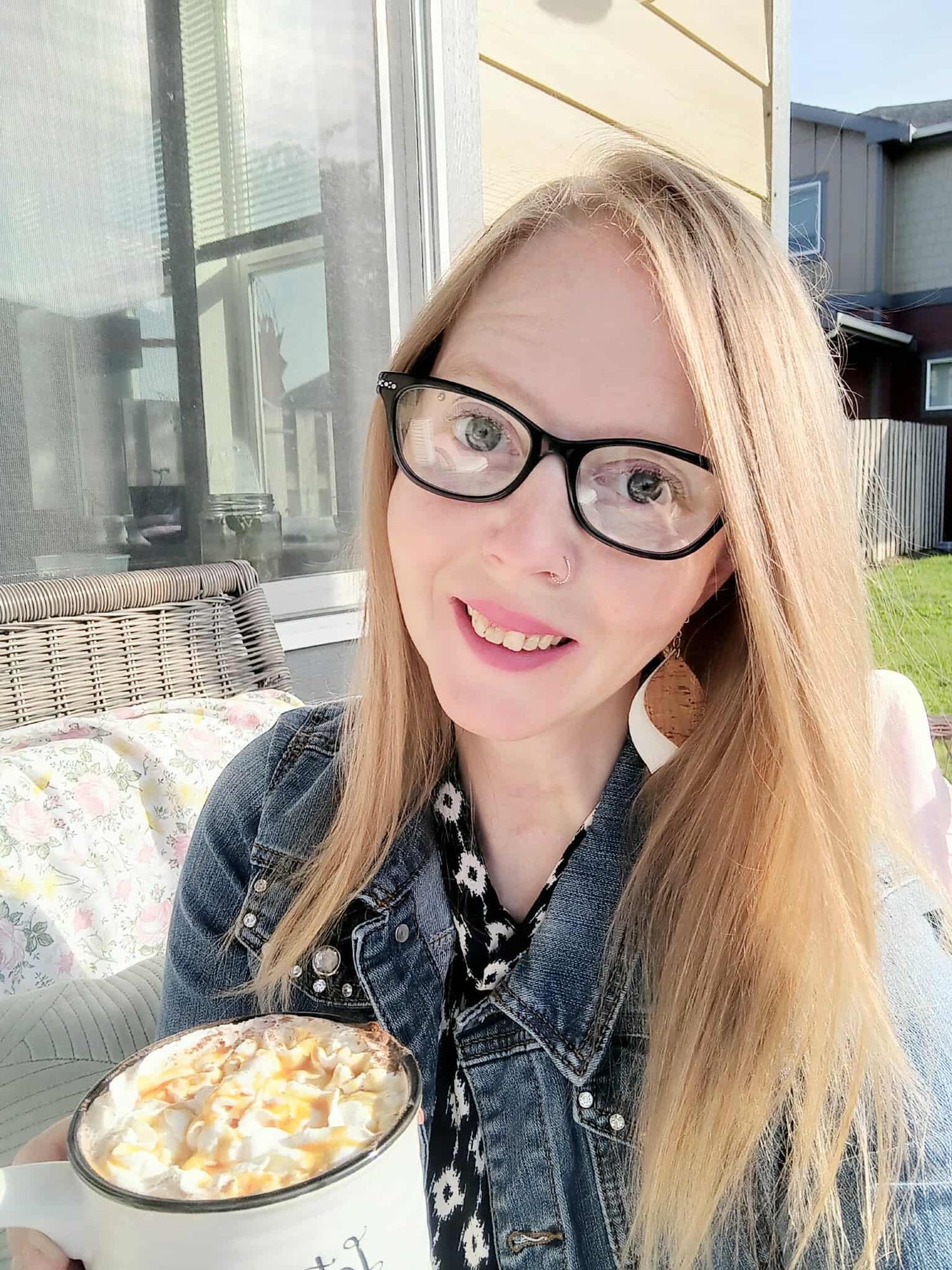 Woman with glasses taking selfie outdoors with a cup of coffee and wearing denim jacket.