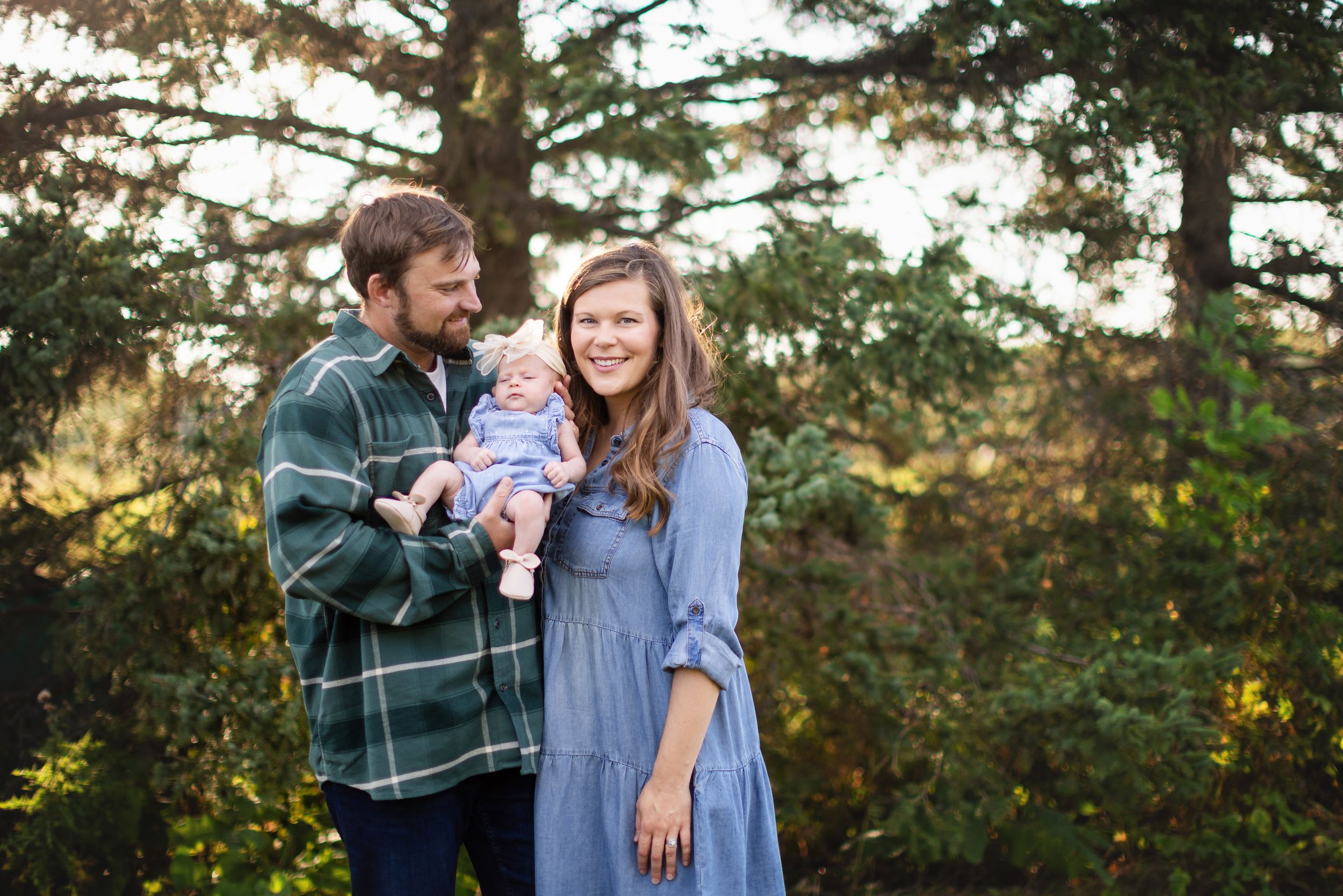 A couple stands together with their baby in a wooded outdoor setting during golden hour lighting.