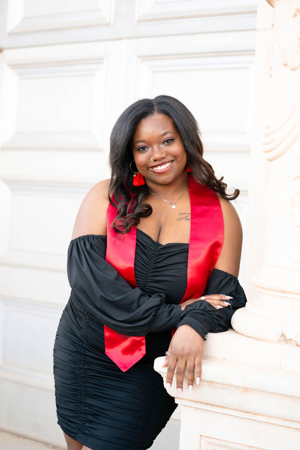 Graduation portrait in black dress with red sash against white architectural backdrop.
