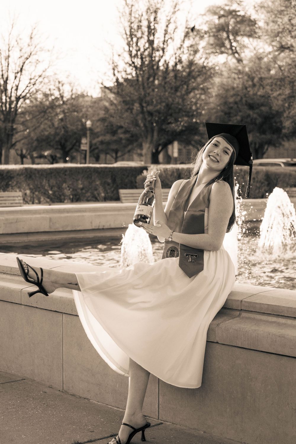 Graduate in white dress and graduation cap poses joyfully by fountain in black and white photo.