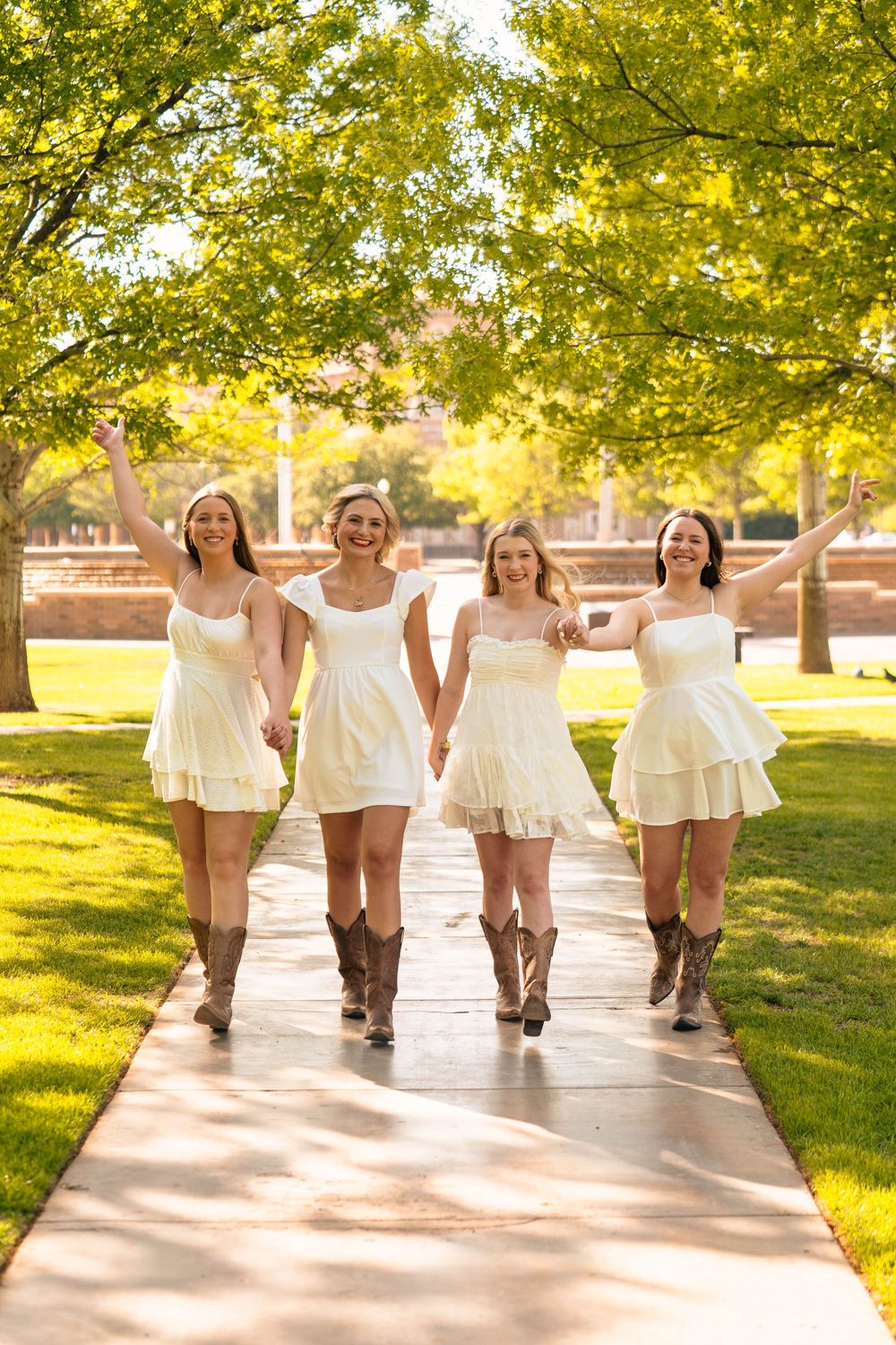 Four friends in white dresses and cowboy boots walk together on campus pathway under trees.