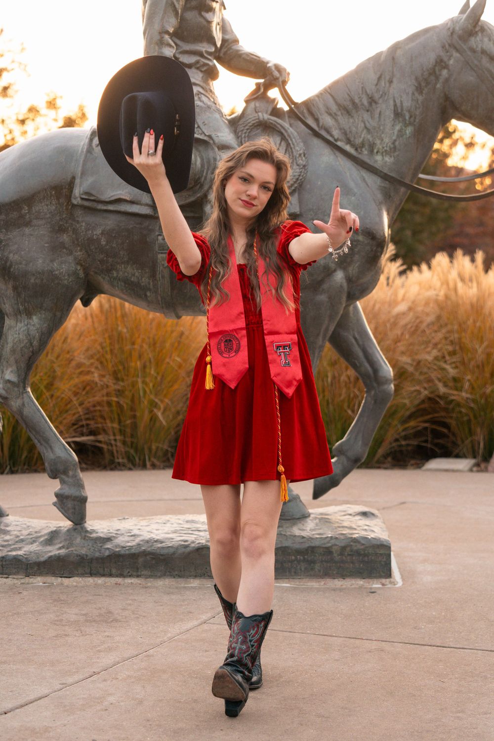 Woman in red dress holds riding helmet while posing by bronze horse statue at sunset.