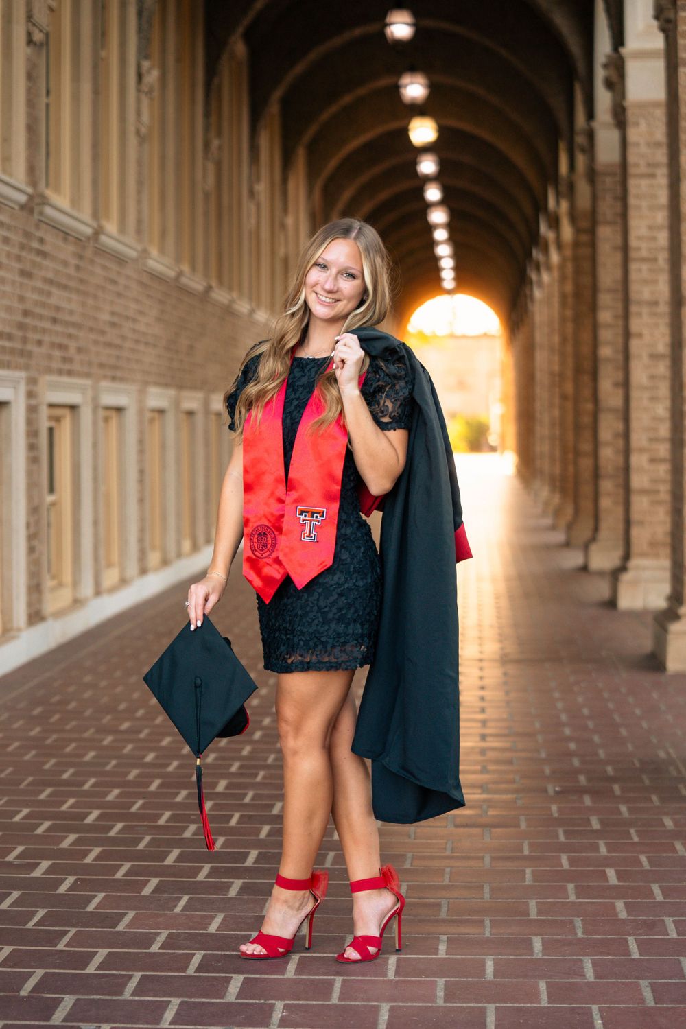 Graduate poses playfully in a historic brick archway wearing red and black academic regalia.
