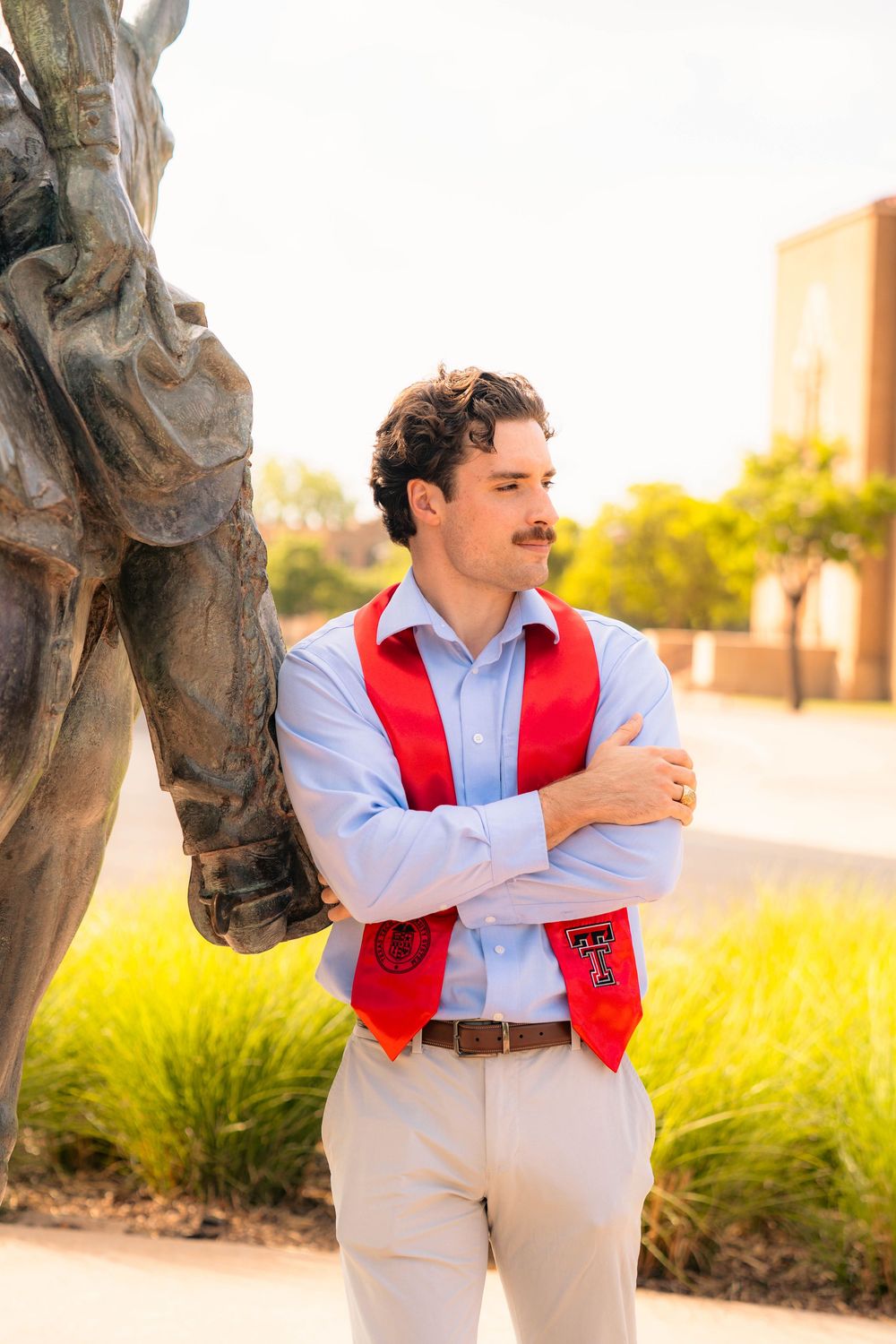 Graduate student in light blue shirt and red vest stands confidently by stone sculpture.