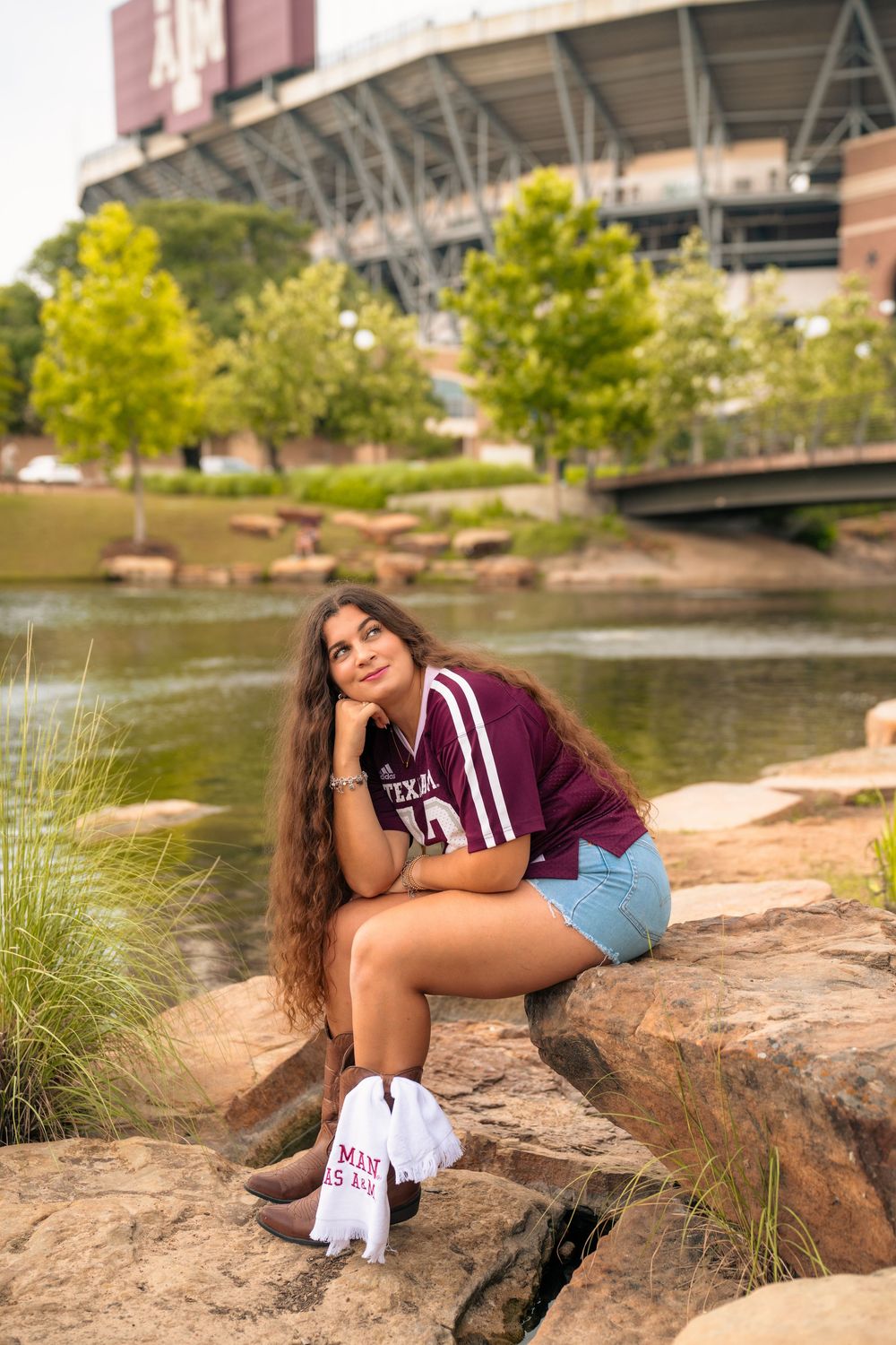 Student sits by a tranquil pond near campus wearing Texas A&M apparel with a stadium visible in the background.