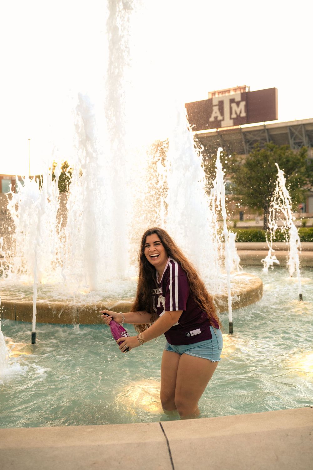 Person splashes in a campus fountain wearing maroon Texas A&M shirt on a sunny afternoon.