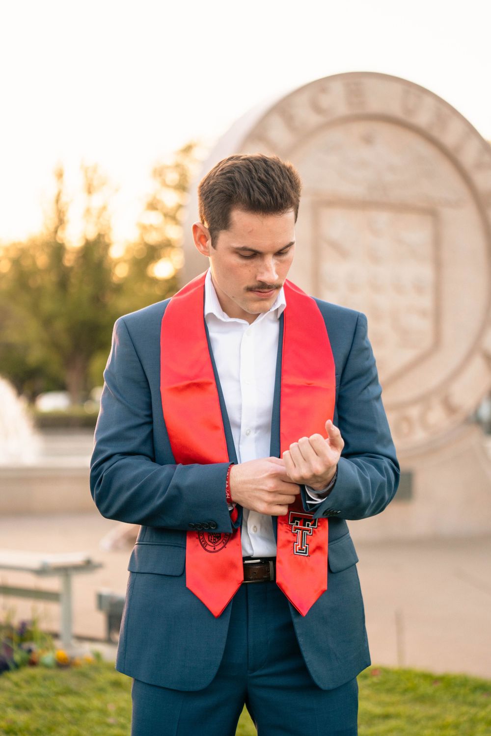 Graduate in navy suit with red stole adjusting sleeve cuff during outdoor graduation photoshoot at sunset.