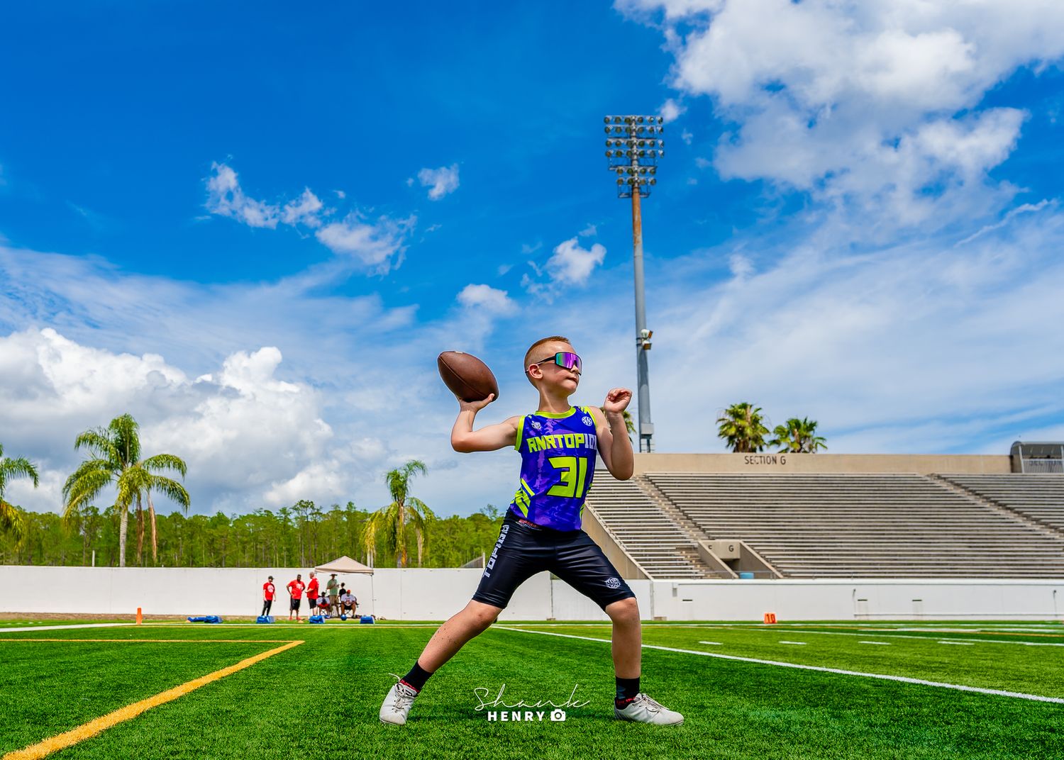 Football player in purple jersey practices throwing form on sunny field with stadium lights.