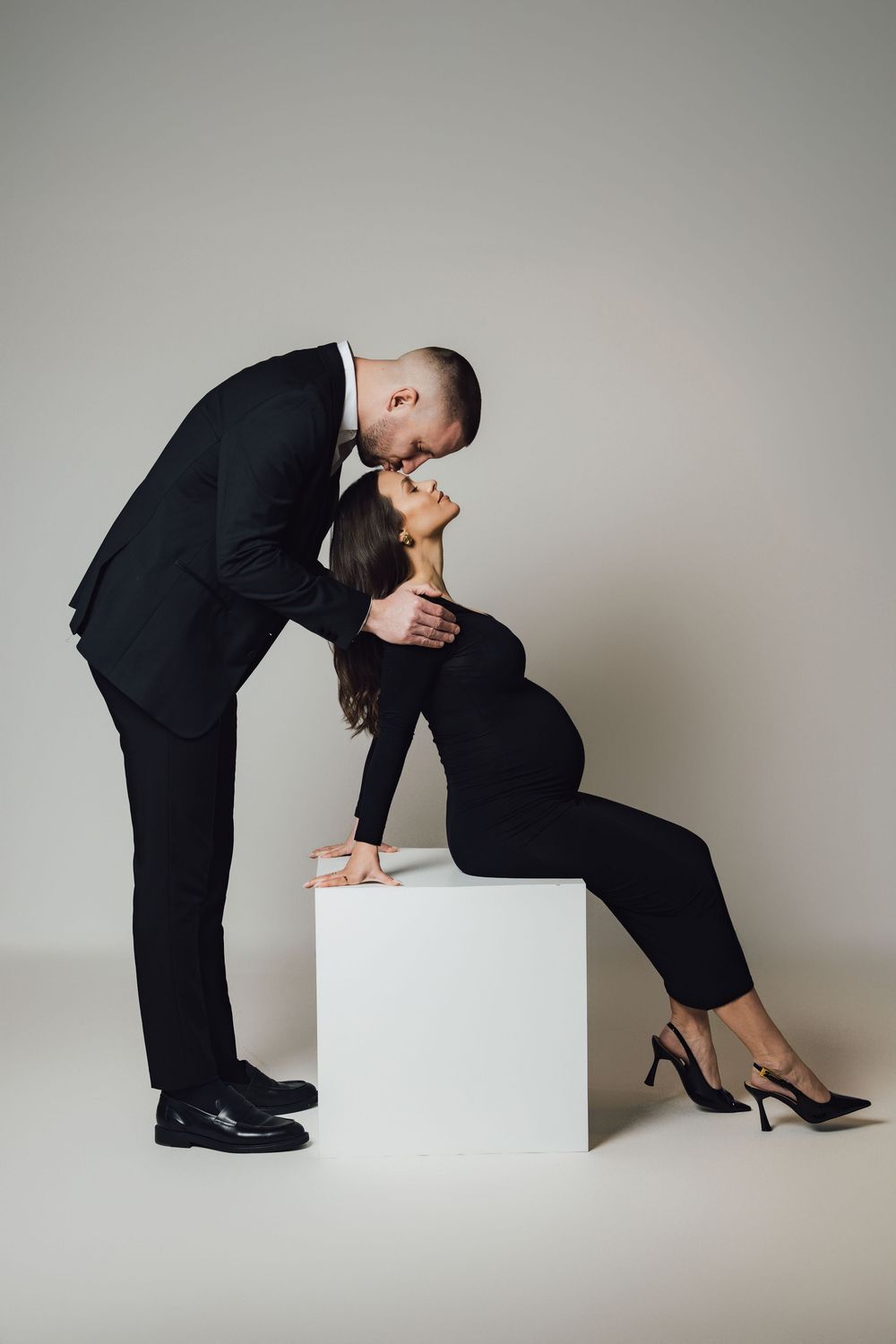 Stylish couple in black formal wear share intimate moment near white cube.