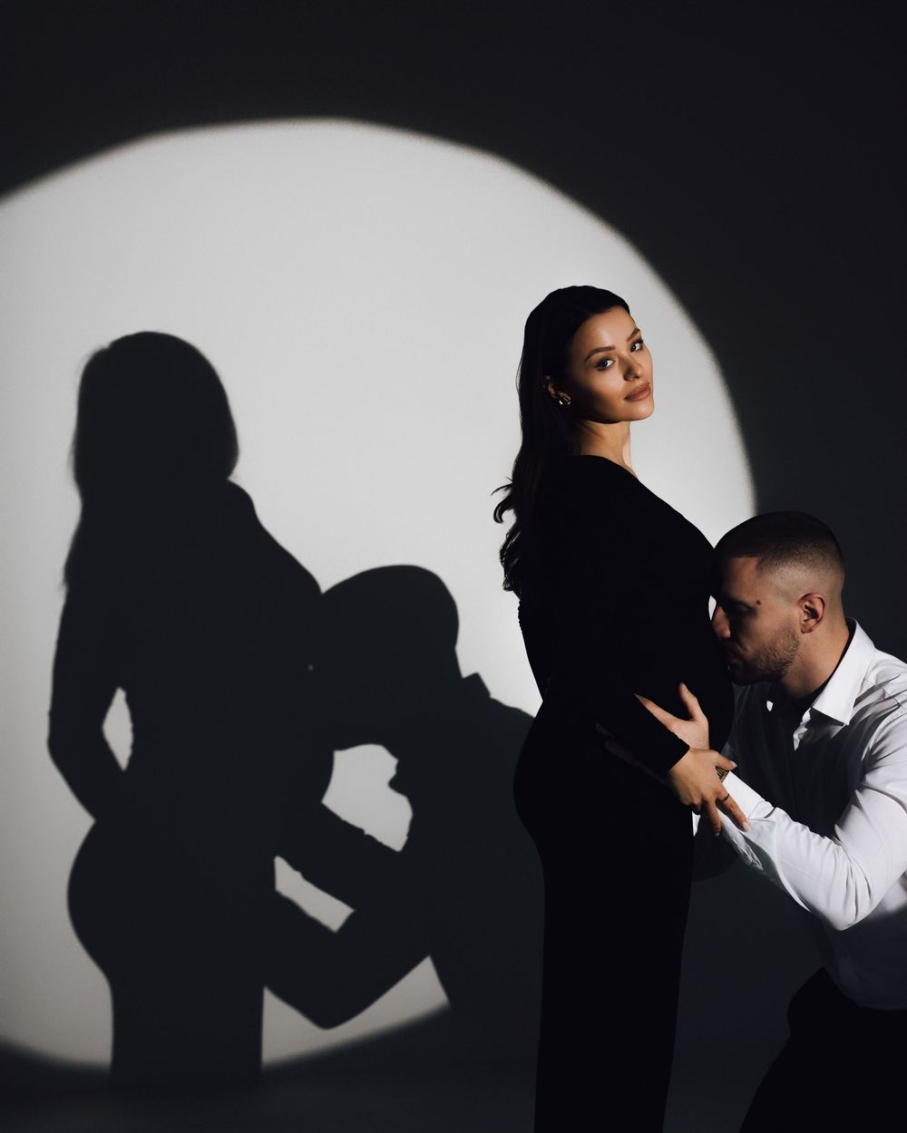 Dramatic maternity photo with couple and shadow projection on white circular backdrop.