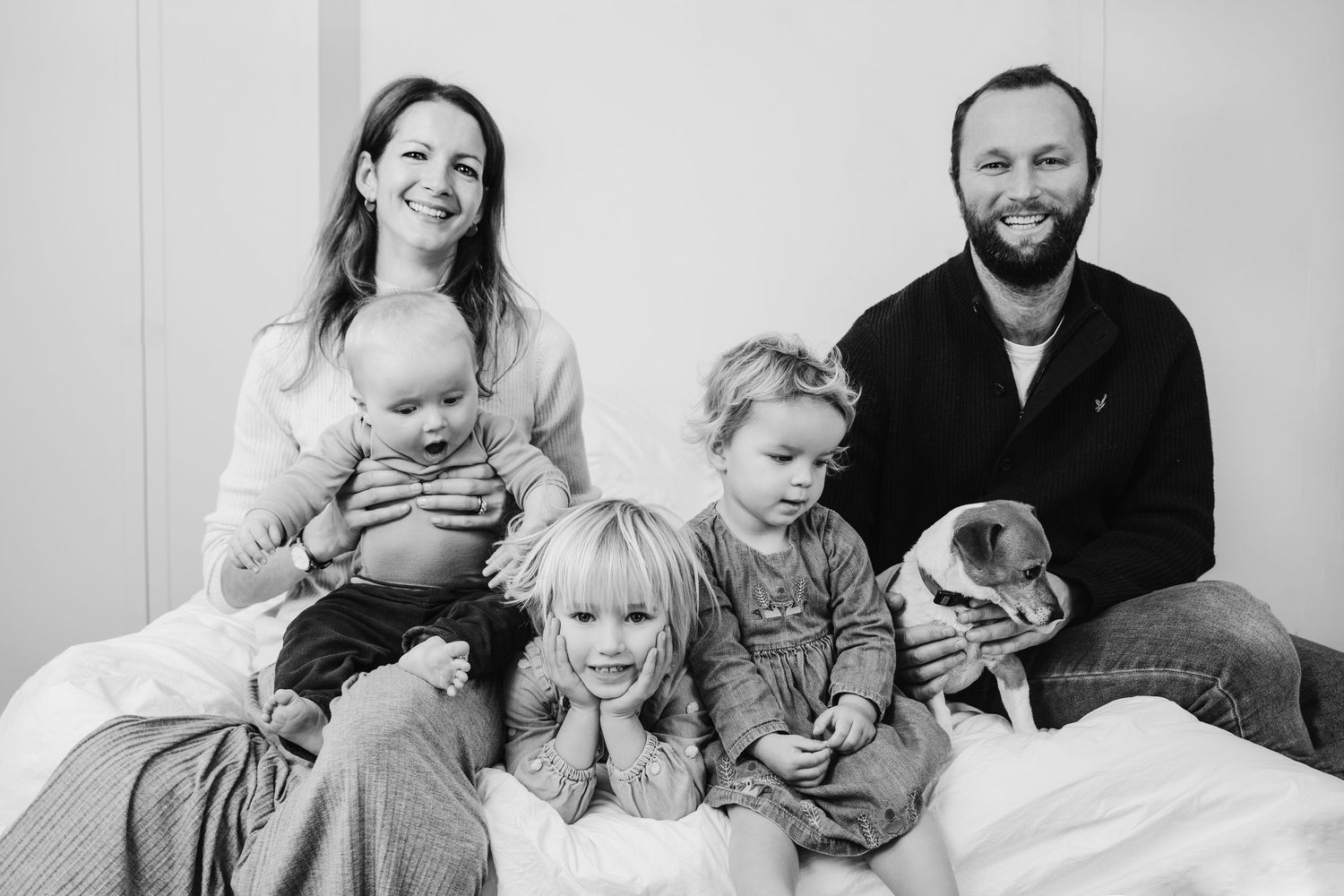 Black and white family portrait with parents, three young children, and a pug sitting together on a bed, shot in Stoke Newington, Hackney.