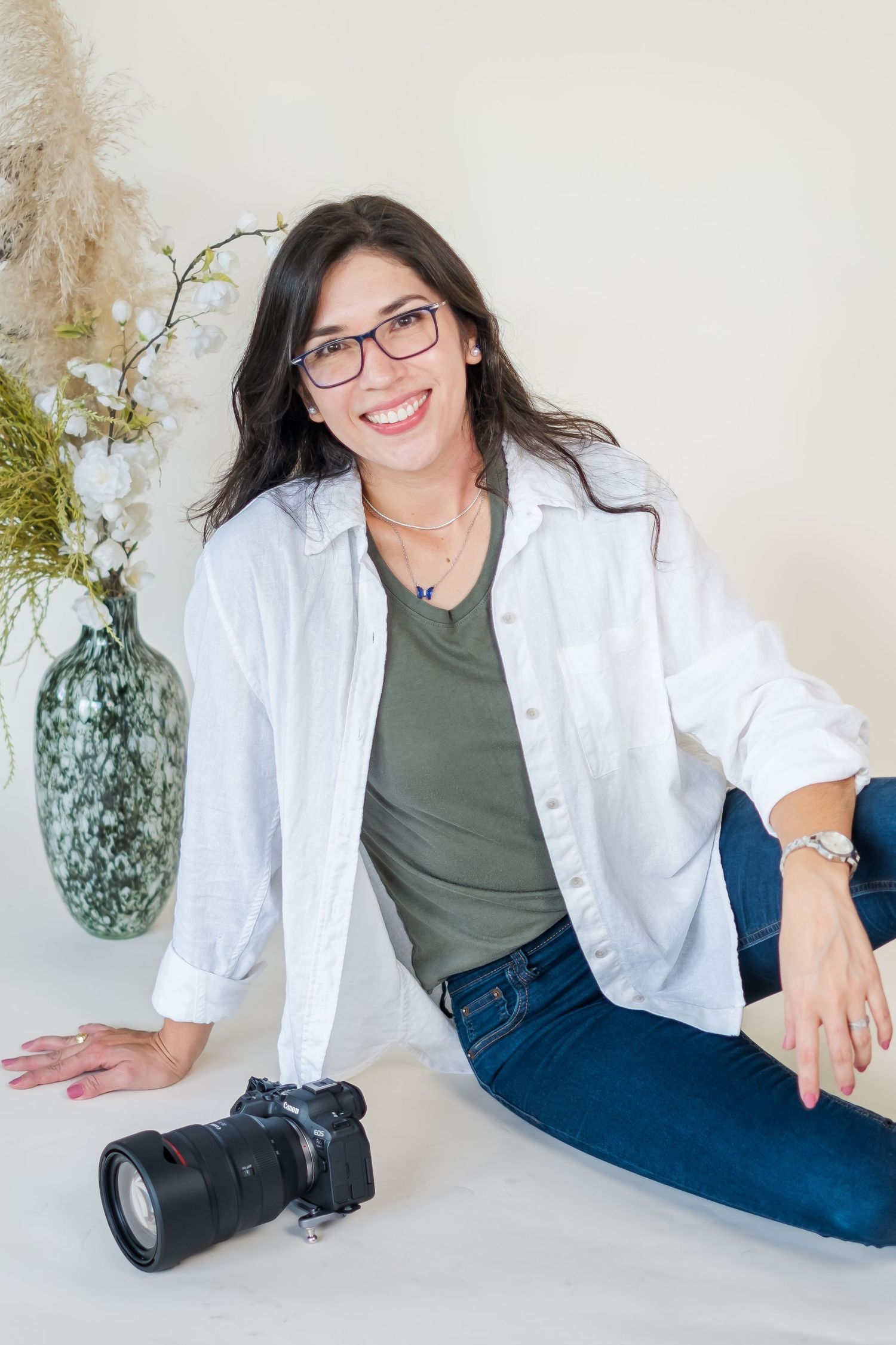 Photographer wearing glasses shows camera while sitting casually in studio setting.