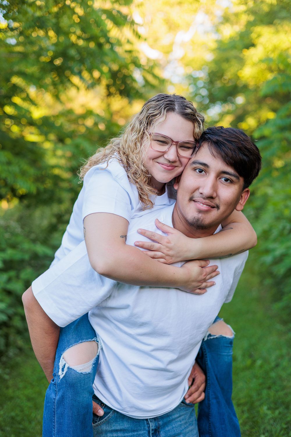 A young couple in white shirts and jeans share a piggyback ride on a green tree-lined path.