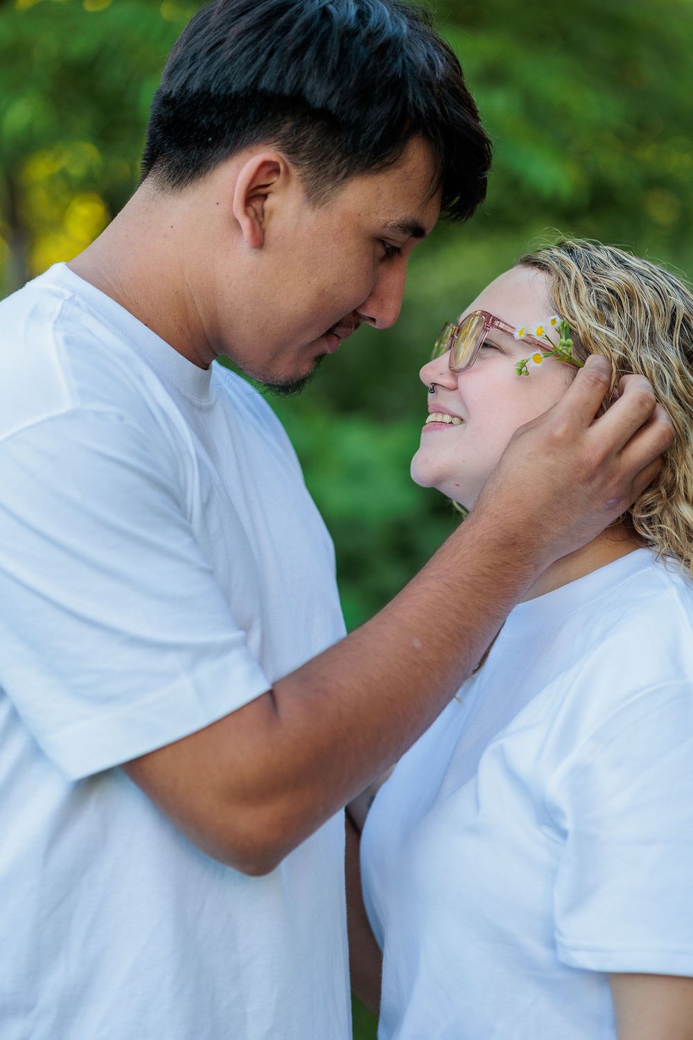Couple in matching white t-shirts share a tender moment outdoors, man gently touching woman's face as she smiles up at him.