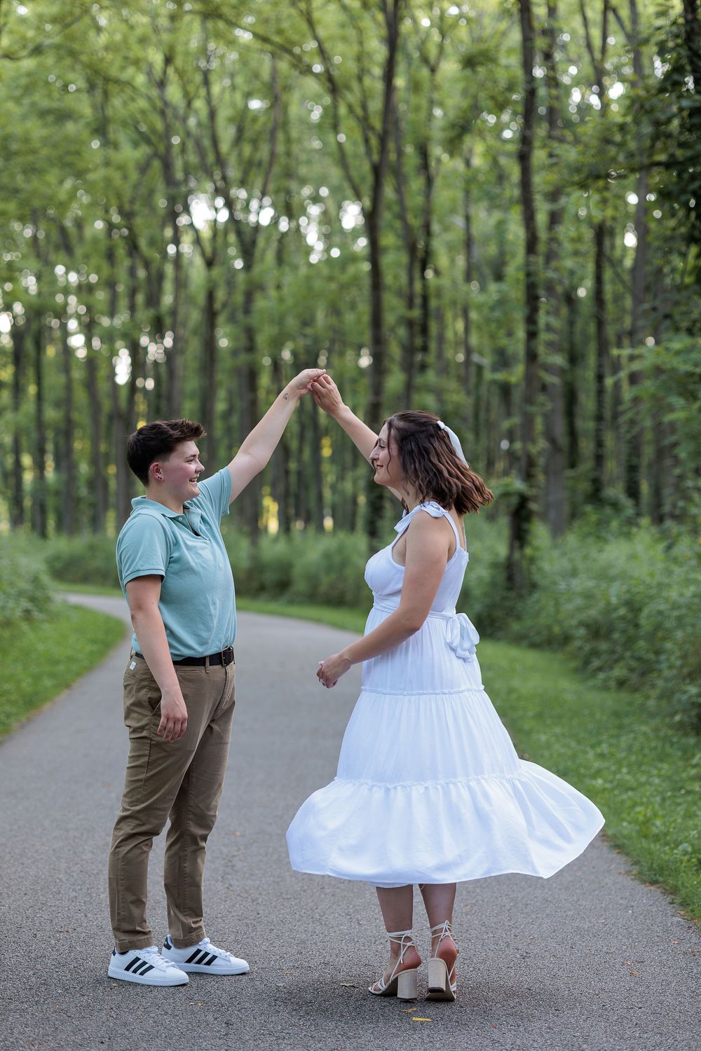 Couple dancing joyfully on a tree-lined path, woman in white dress being twirled by partner in teal shirt.