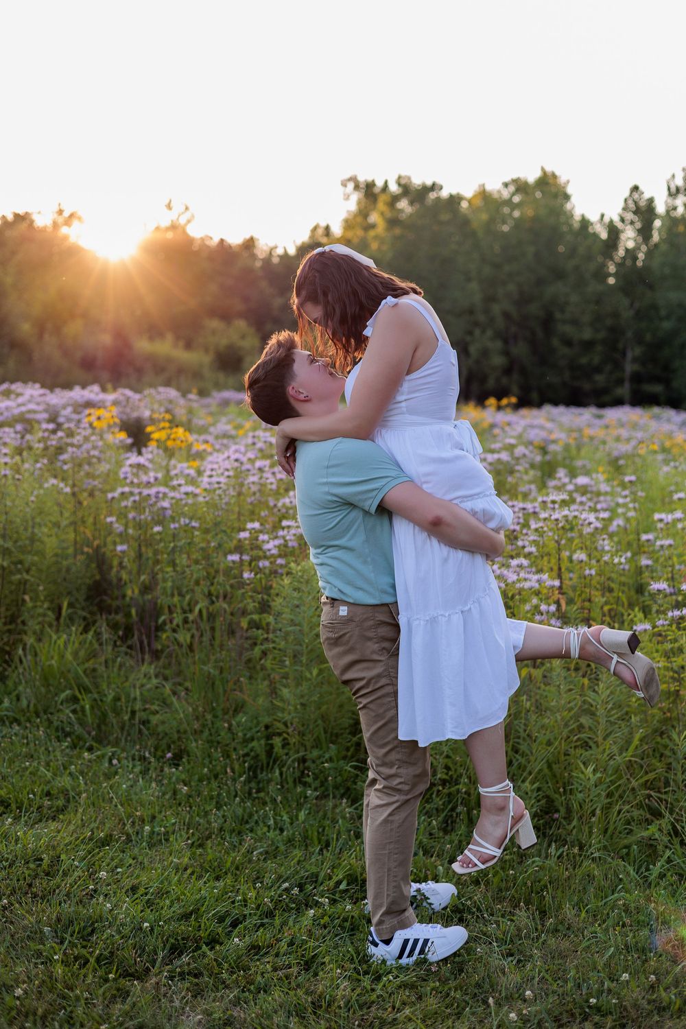 Couple sharing a romantic kiss in a wildflower field at golden sunset, man lifting woman in white dress.