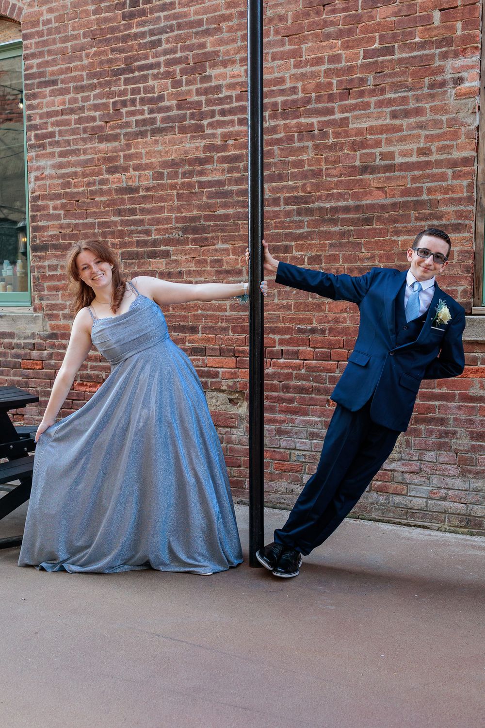 A young couple in prom attire pose by a pole against a brick wall, wearing a blue gown and navy suit.