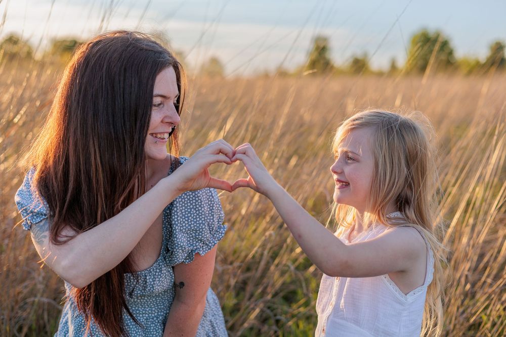 A mother and young daughter make a heart shape with their hands in a golden field at sunset.