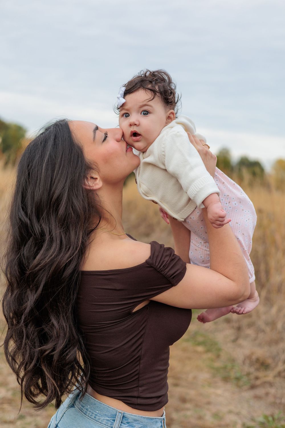 A young mother with long dark hair kisses her surprised baby outdoors in a golden field.