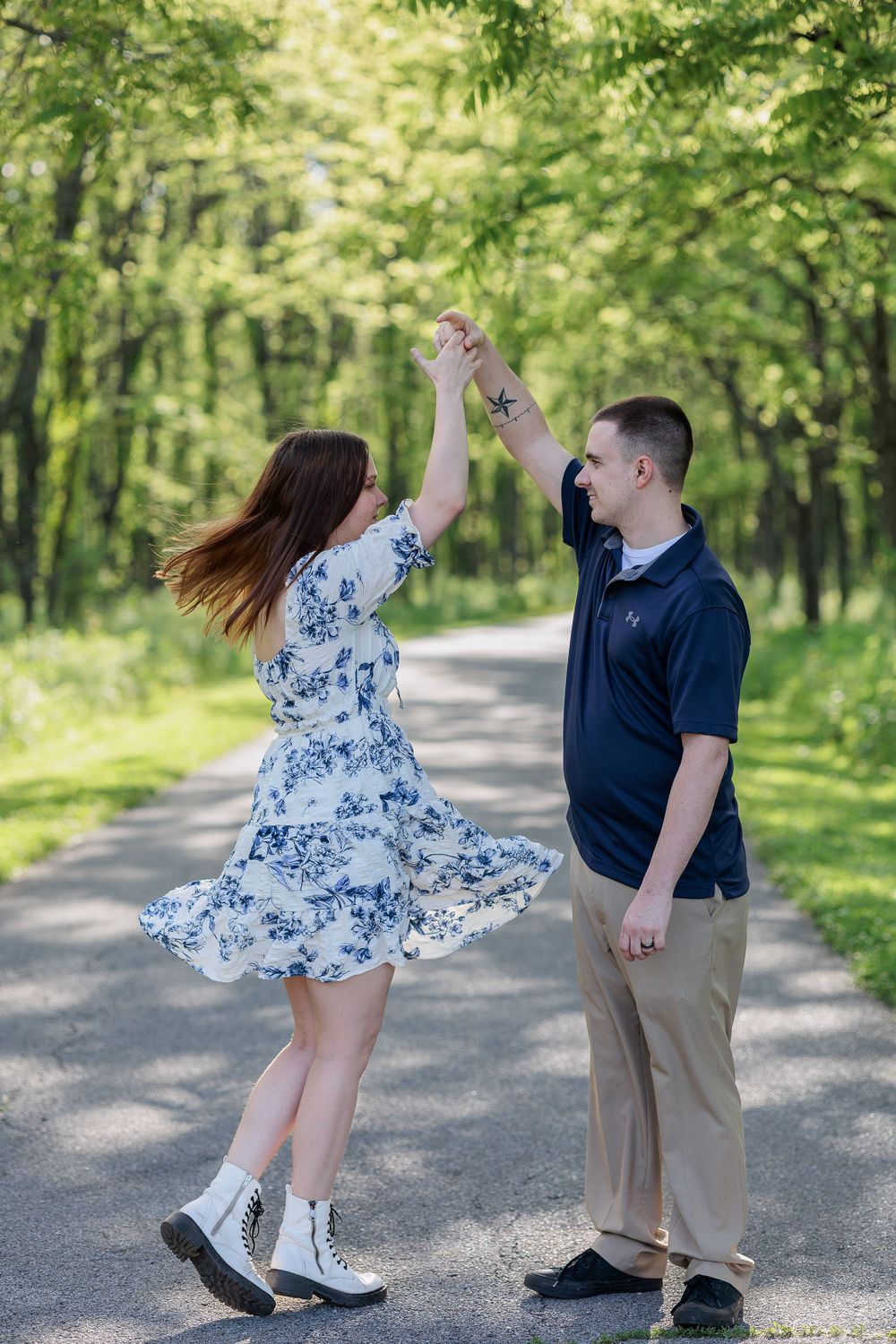 A couple dances joyfully on a tree-lined path, the woman spinning in a floral dress while holding her partner's hand.