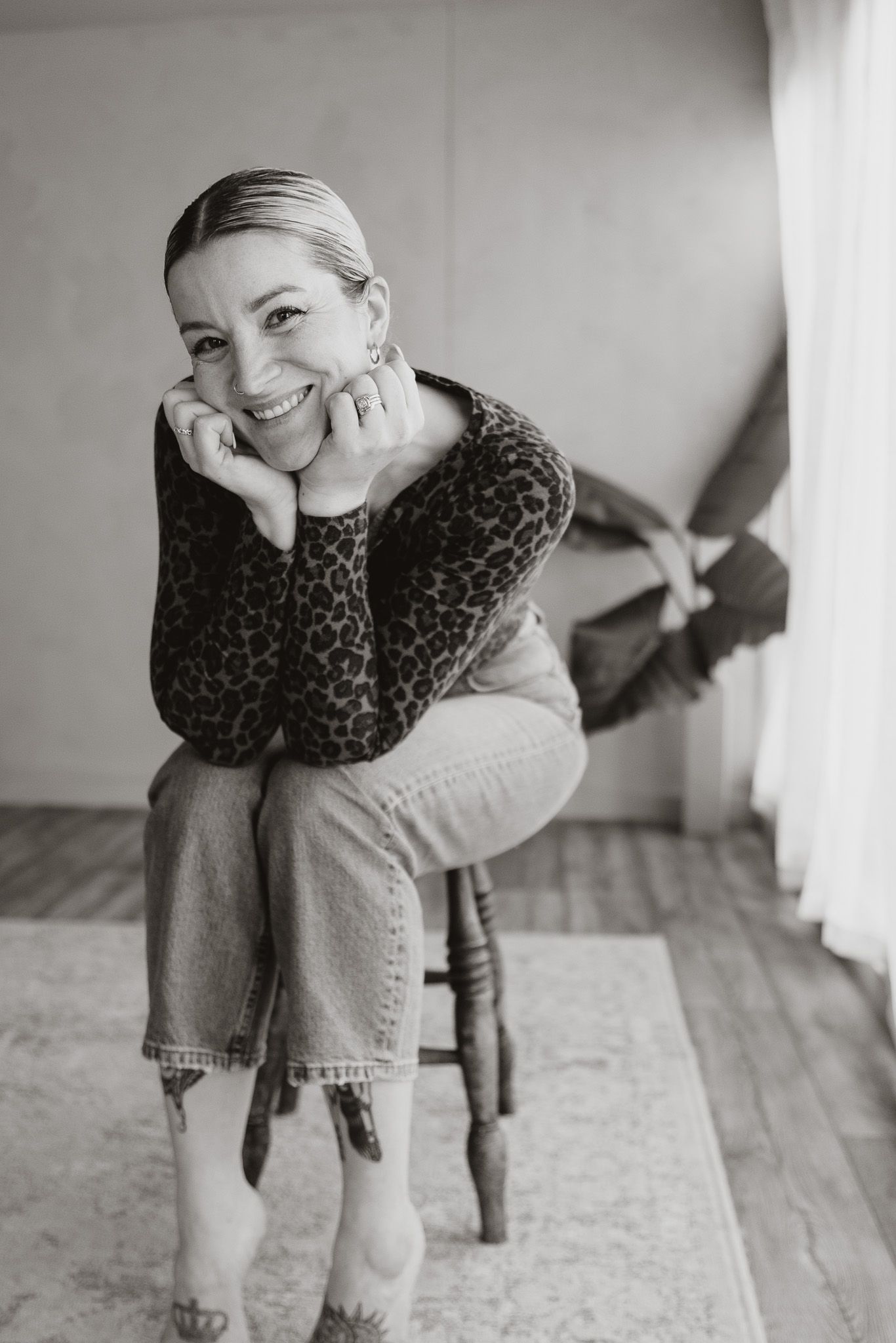 Black and white photo of Vix in jeans and a leopard print top sitting on a wooden chair in her studio.