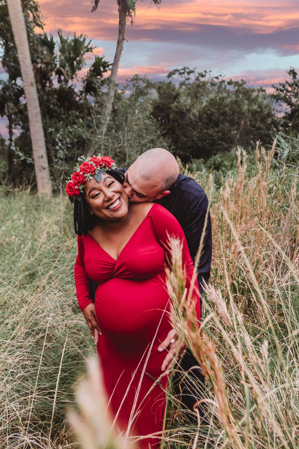 Pregnant woman in red dress with floral crown smiles as partner kisses her cheek at sunset in a grassy field at Riverbend Park Jupiter.