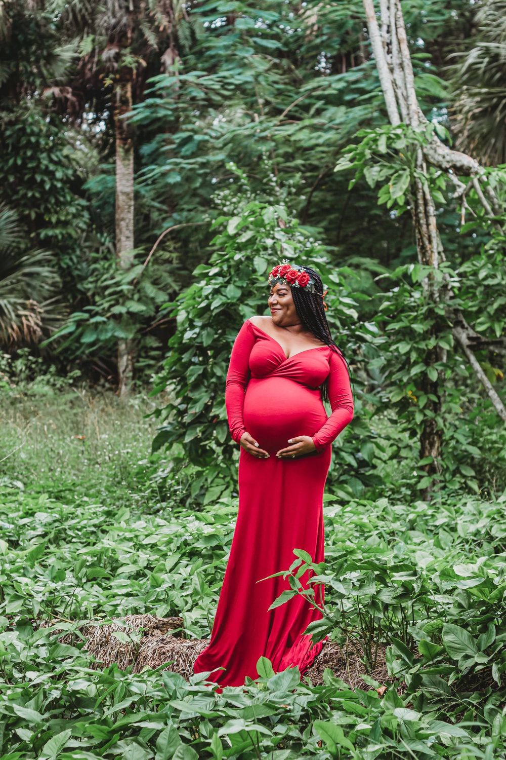 Smiling pregnant woman in red gown and floral crown cradling baby bump in lush green tropical forest setting  at Riverbend Park Jupiter.