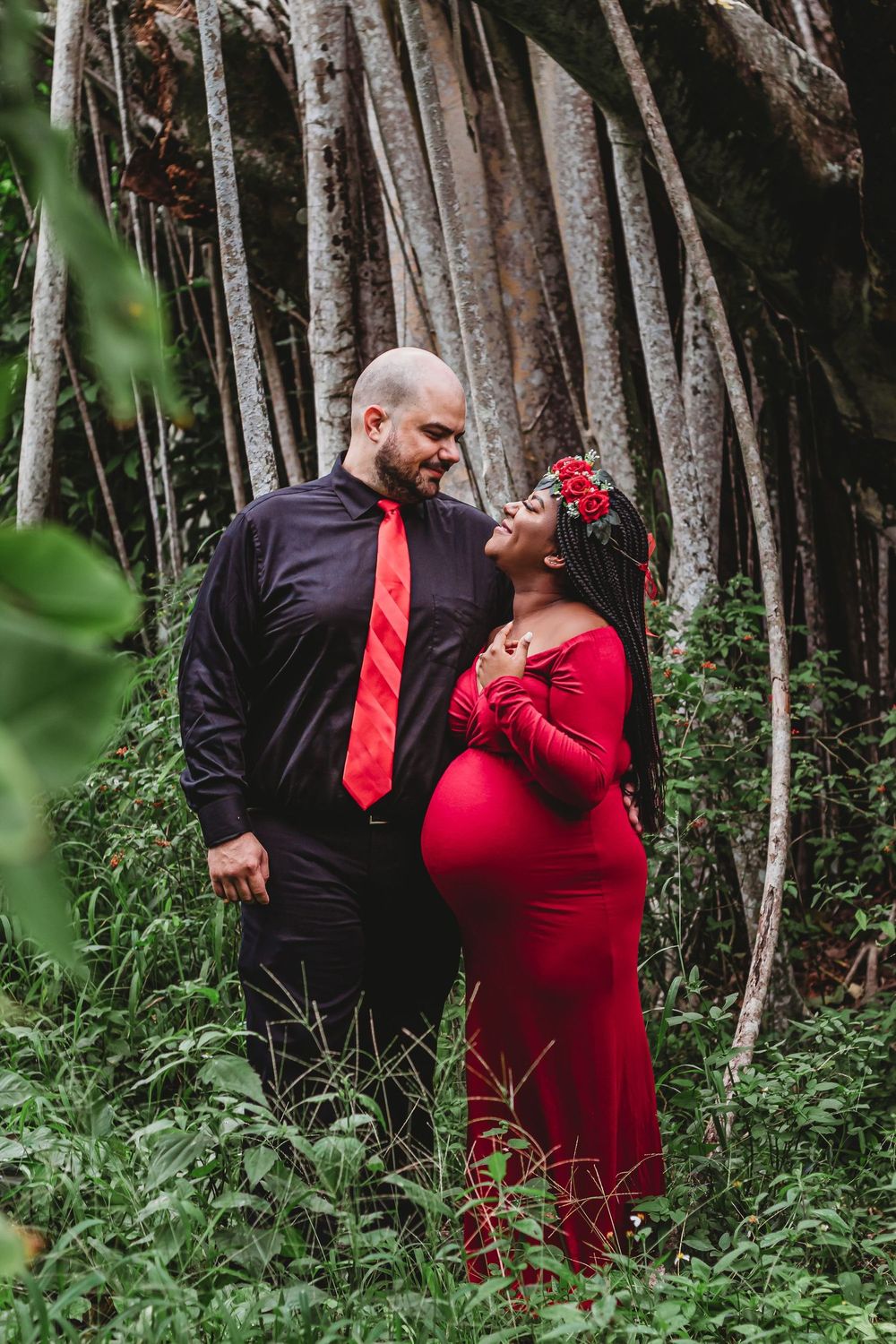 Pregnant couple in matching red and black outfits posing romantically in a lush green forest setting at Riverbend Park Jupiter.