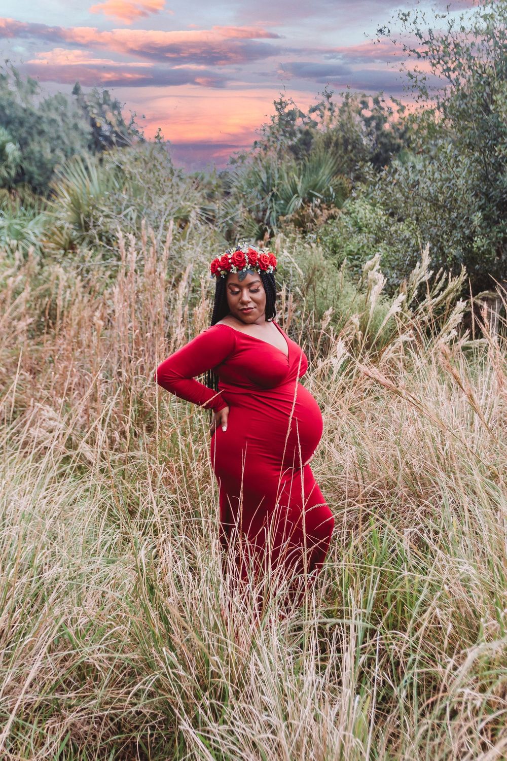 Pregnant woman in red gown and floral crown stands in tall grass at sunset at Riverbend Park Jupiter.