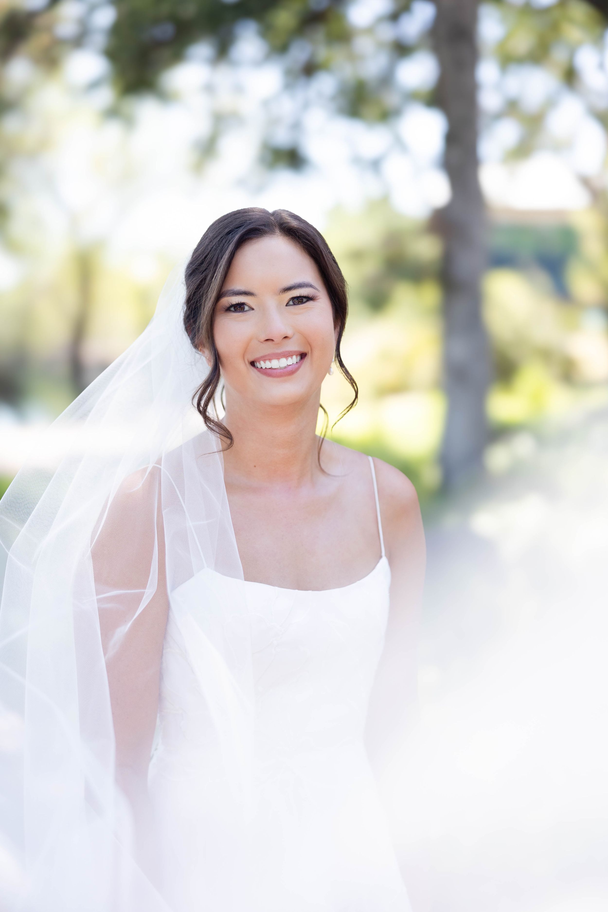 Bride with veil swooping towards camera on sidewalk with trees behind them at Lakeside Pavilion in Chico, California
