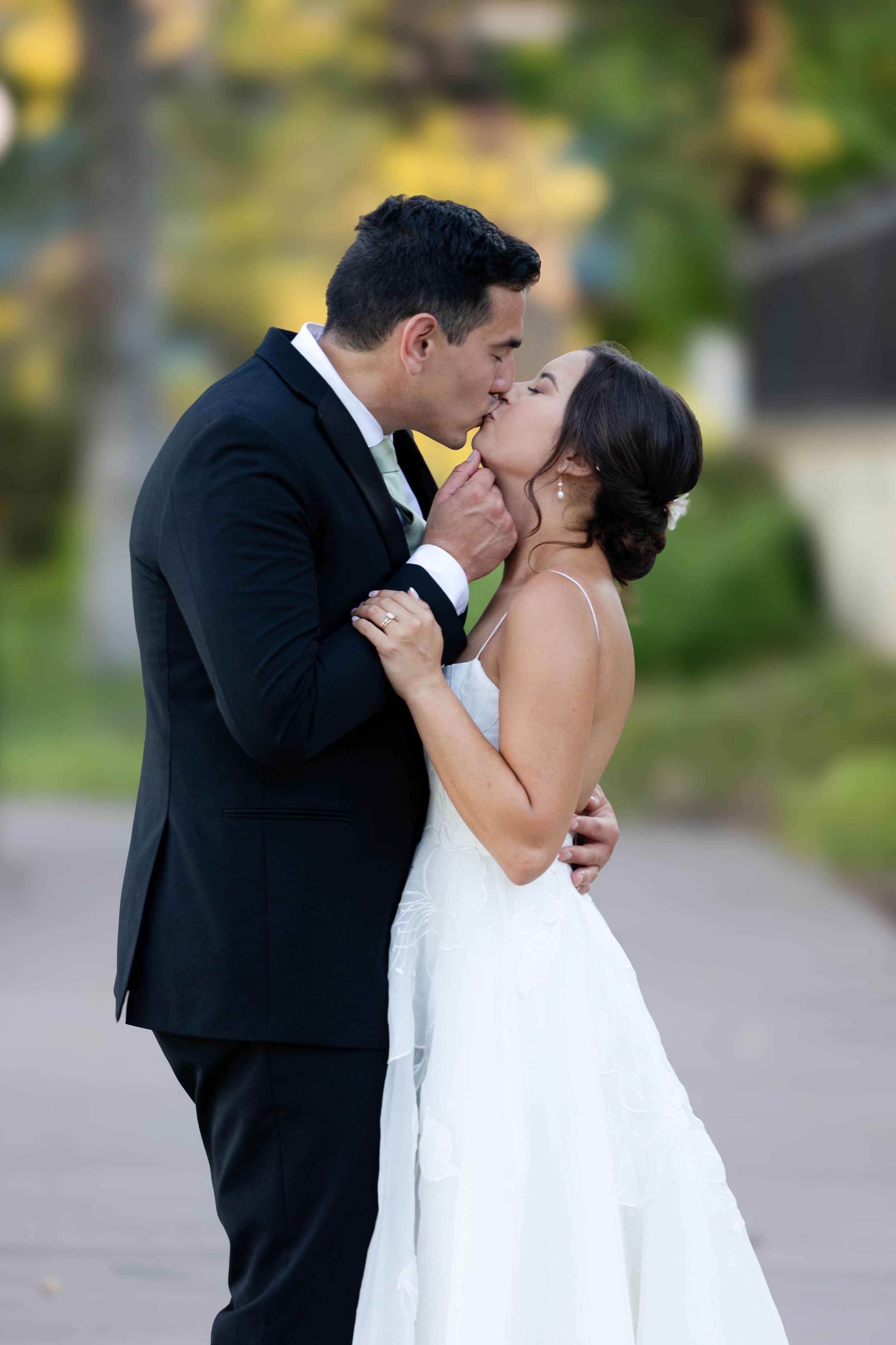 Bride and and Groom Kissing on sidewalk with trees behind them at Lakeside Paviillion in Chico, California