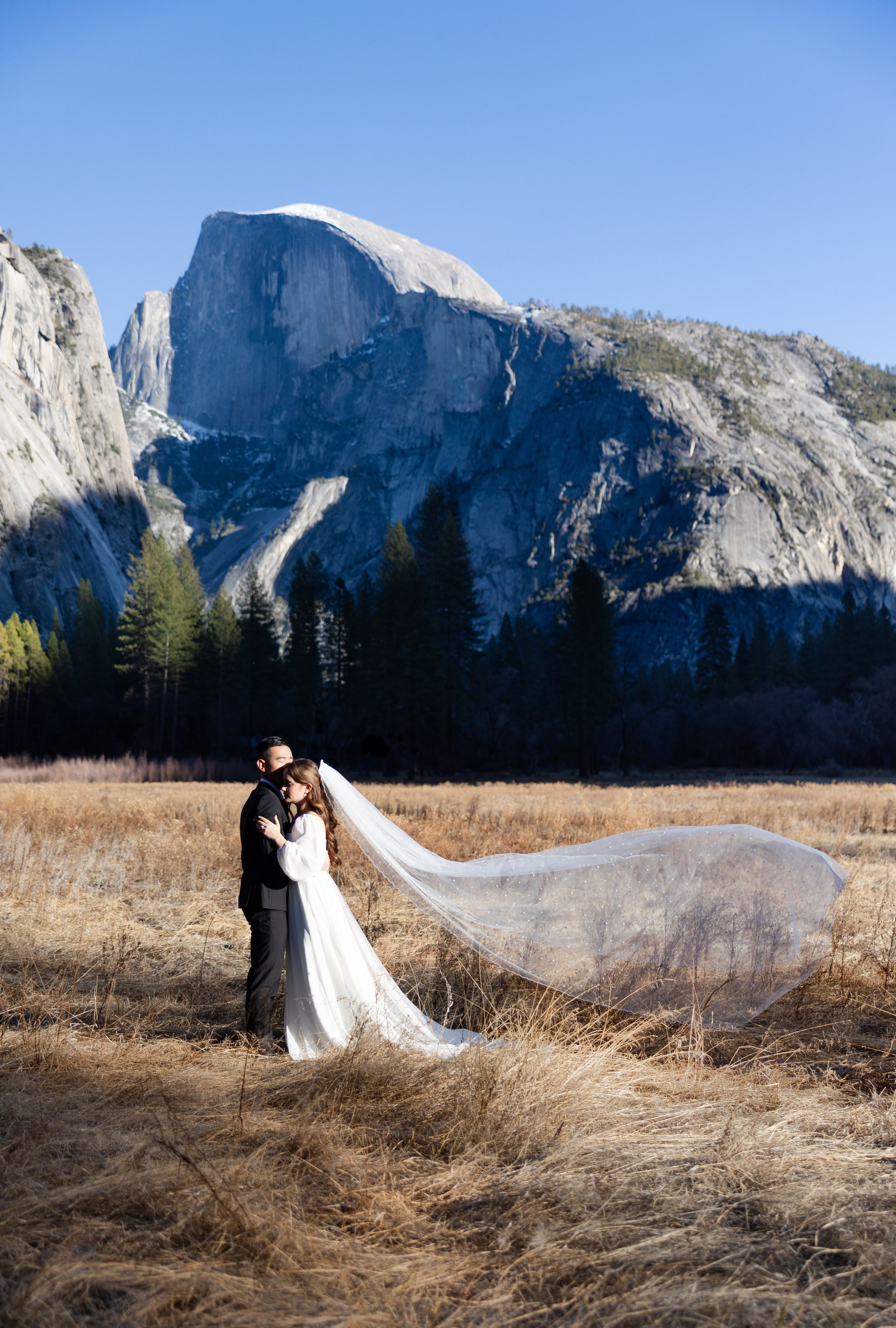 A couple poses romantically in a grassy meadow with dramatic mountain peaks of Yosemite Valley as their backdrop.