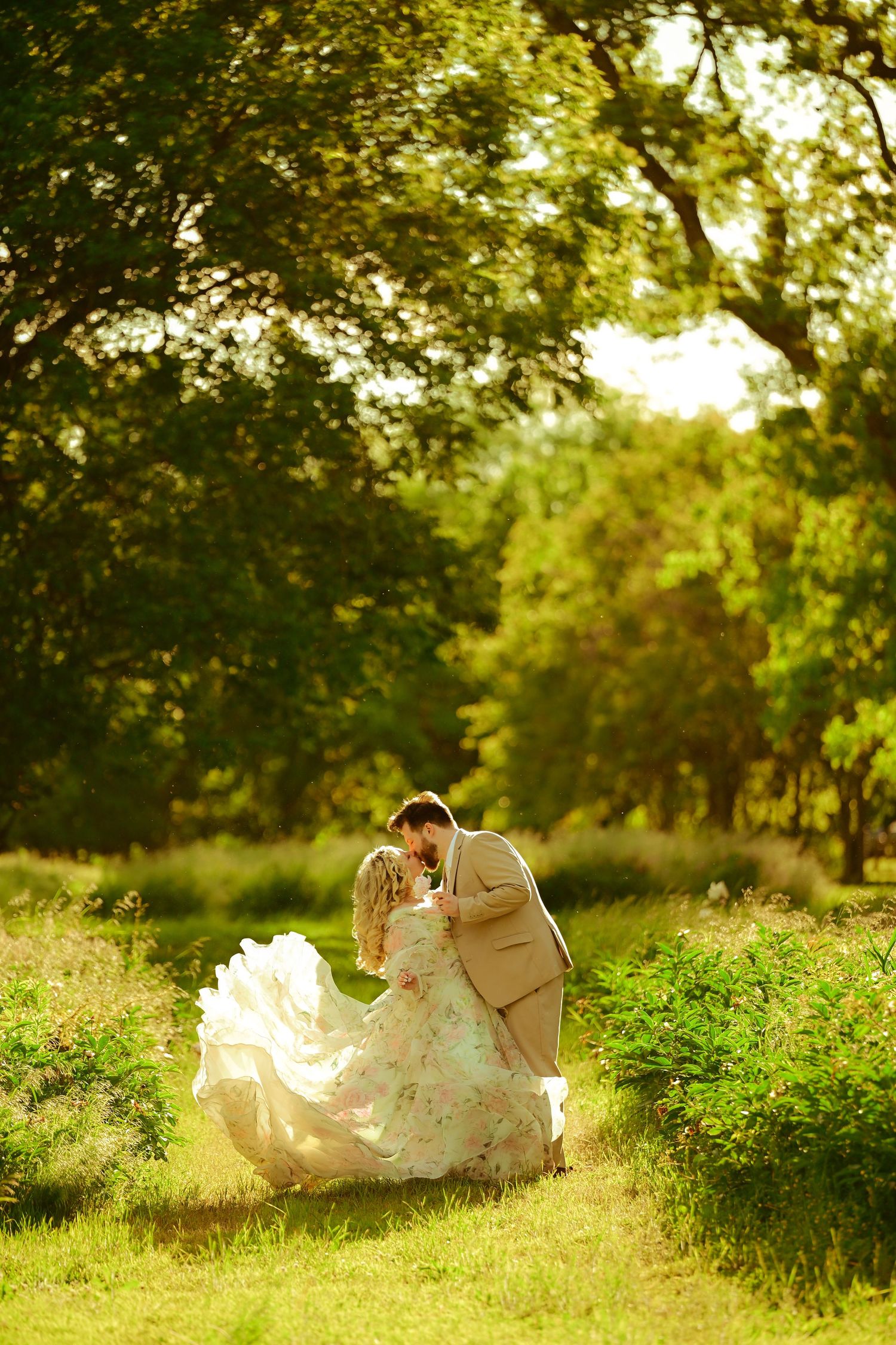 A romantic wedding couple shares a tender moment in a sun-dappled garden setting with flowing white dress details.