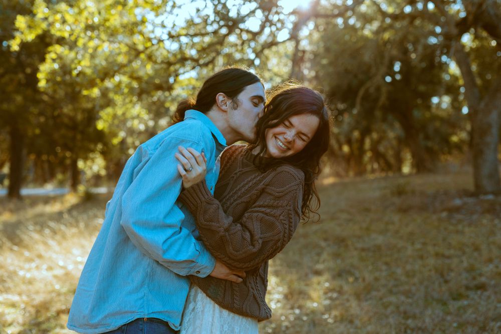 A man kissing a smiling woman on the cheek during a couple's photo shoot in a sun-drenched Central Texas grove.