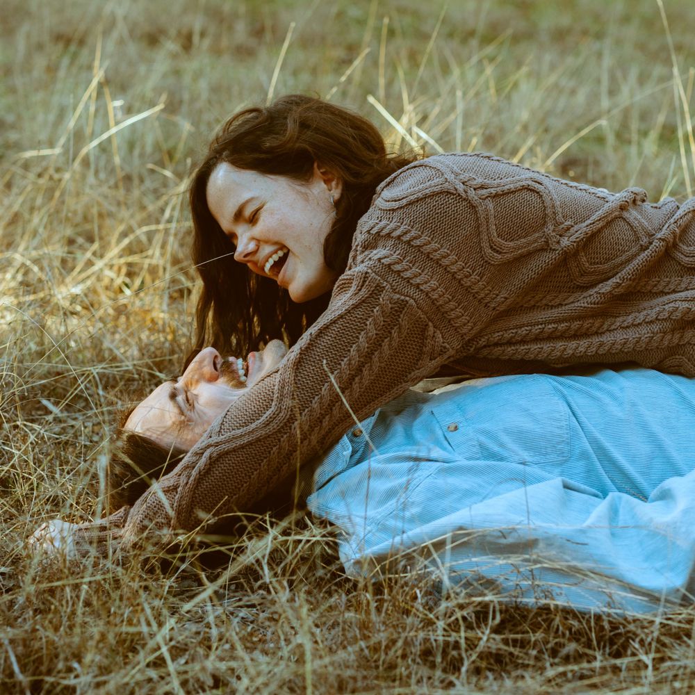A laughing couple playfully laying together in a tall grassy field during a Central Texas portrait session.